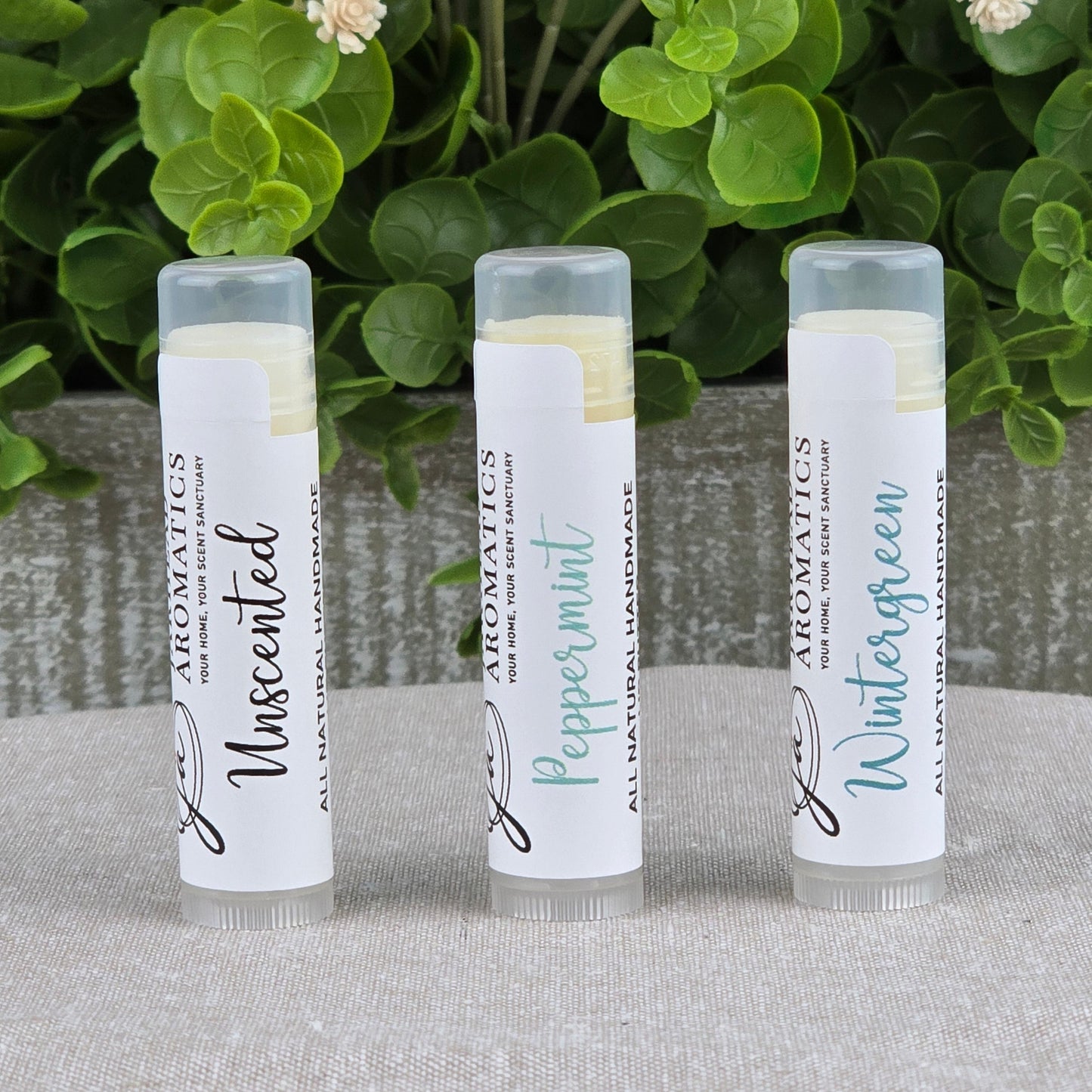 Three upright tubes of Jarhead Aromaticsโ Lip Butter - All Natural in Unscented, Peppermint, and Wintergreen sit on gray cloth, surrounded by green leaves and cream flowers, highlighting their ultra-nourishing natural formula.