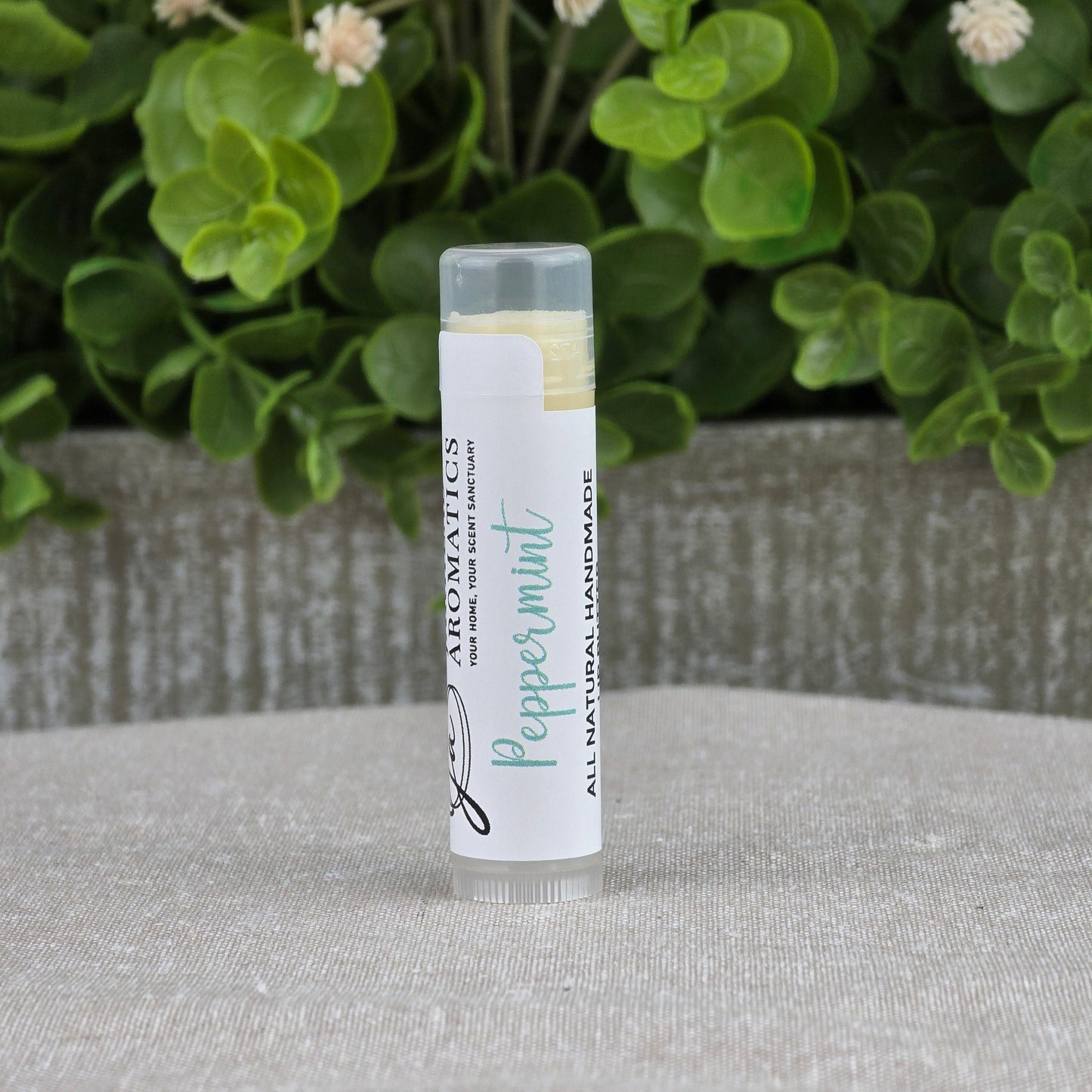 A tube of Jarhead Aromatics Lip Butter - All Natural stands upright on a gray fabric surface, with green leaves and small white flowers blurred in the background, suggesting its natural origins.