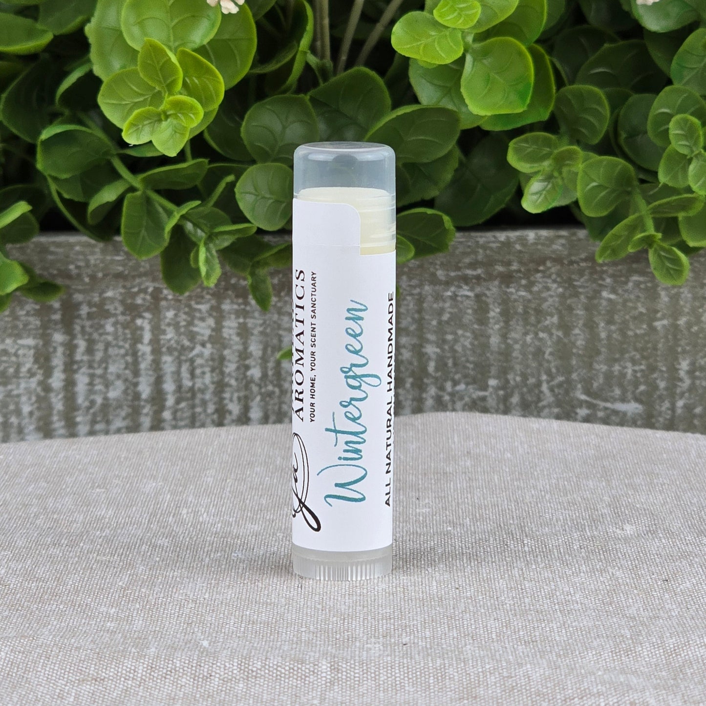 A tube of Jarhead Aromatics Lip Butter - All Natural stands upright on a textured surface, with green leafy plants and small white flowers in the background.