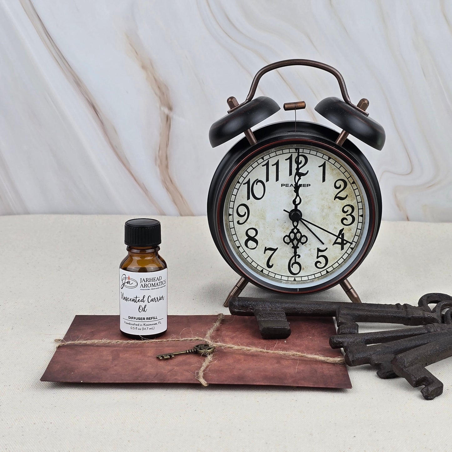 A vintage black alarm clock, an old-fashioned key, two large decorative keys, and a Jarhead Aromatics Unscented Carrier Oil - Cold Air Diffuser Refill rest on a brown envelope tied with twine against a light marble background.
