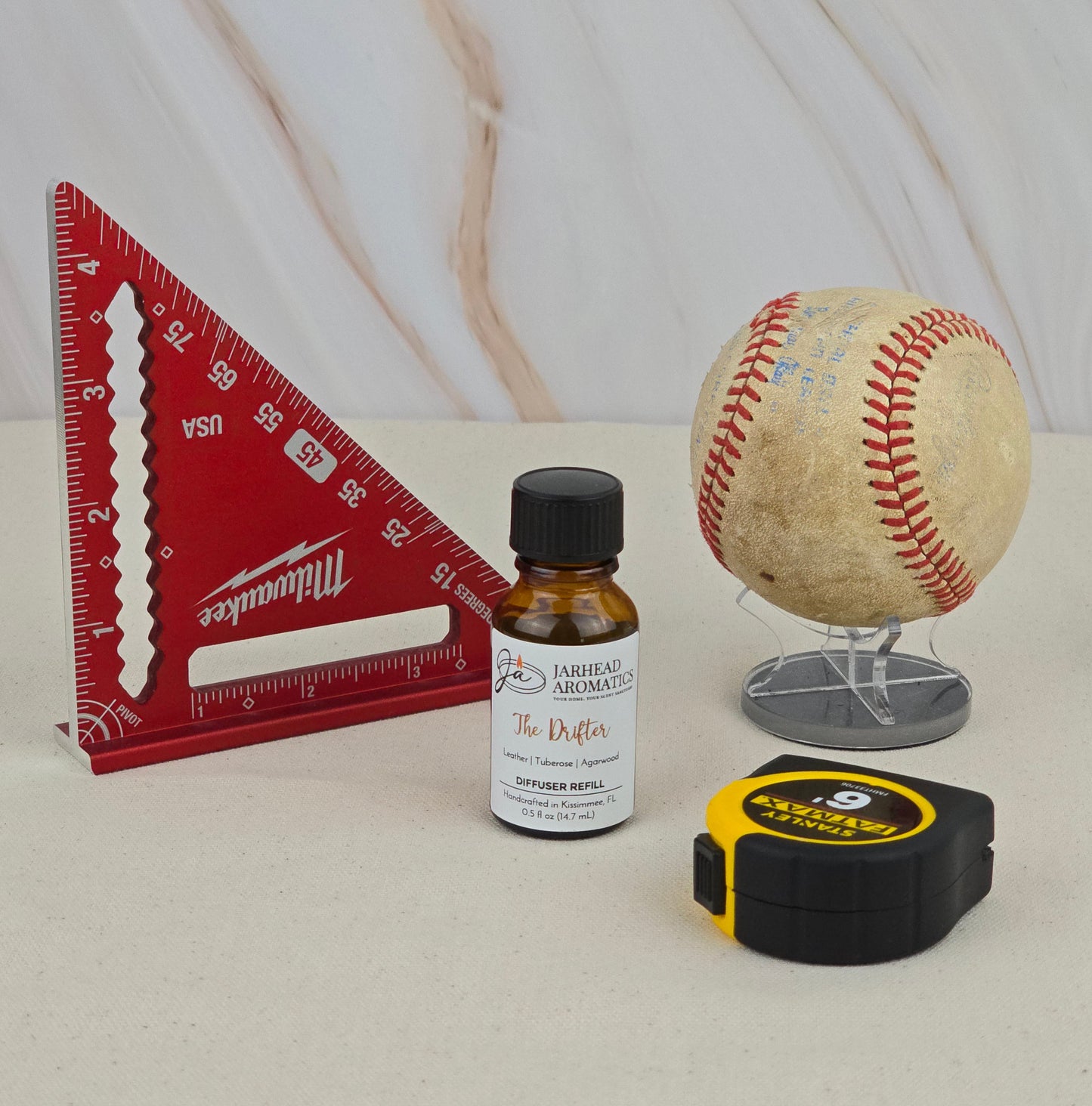 A red triangular ruler, The Drifter - Cold Air Diffuser Refill by Jarhead Aromatics, a worn baseball on a stand, and a yellow tape measure rest on beige against a light marble backdrop.