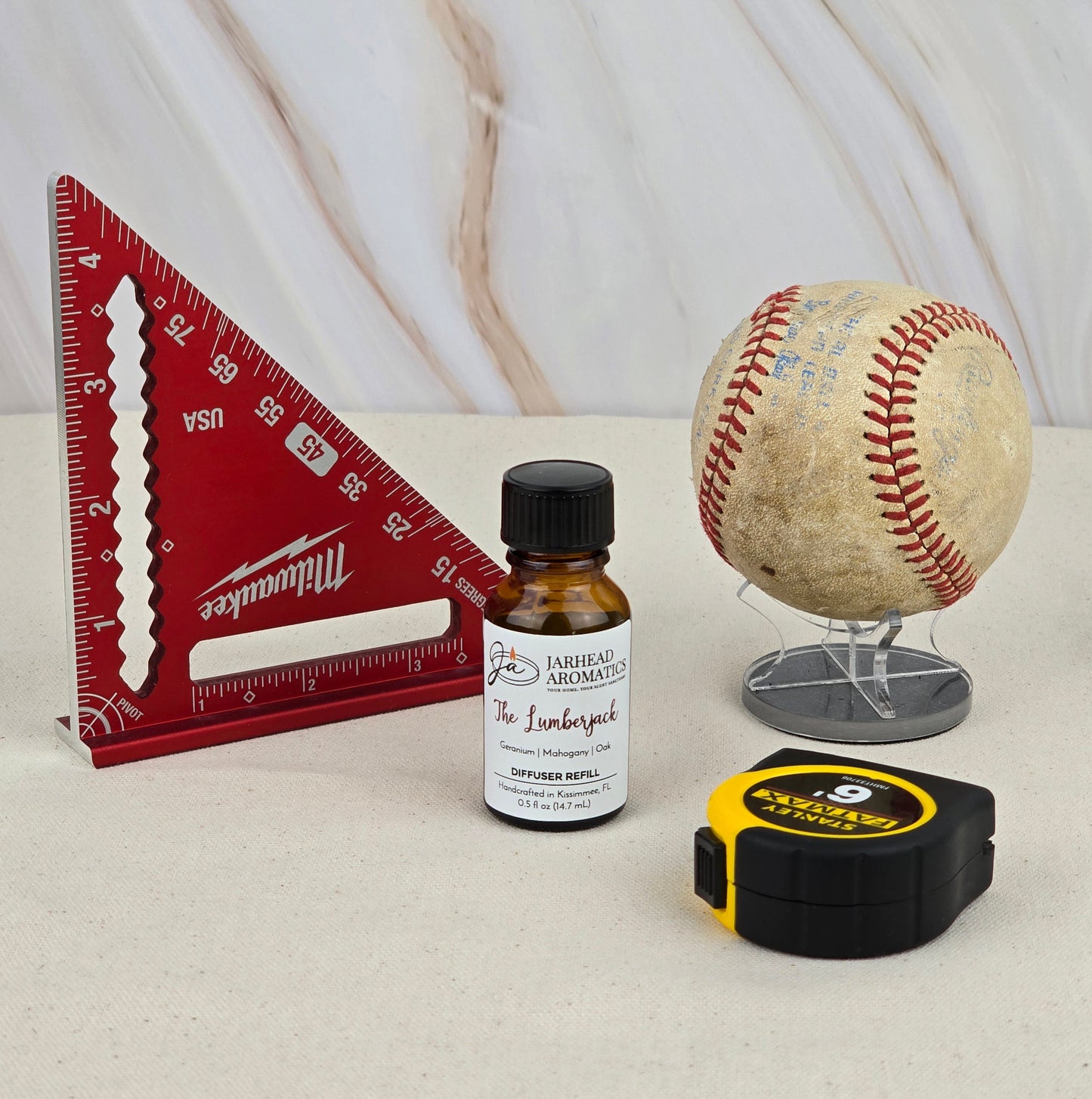 A red triangular ruler, The Lumberjack - Cold Air Diffuser Refill by Jarhead Aromatics, a worn baseball on a stand, and a yellow tape measure rest on a light surface with a marble-patterned background.