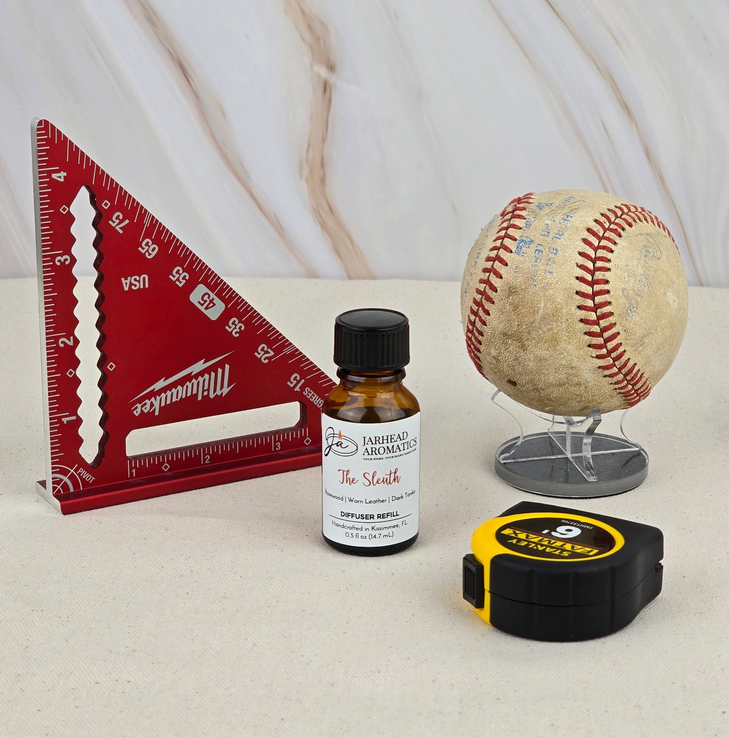 A red triangle ruler, a bottle of Jarhead Aromatics’ The Sleuth – Cold Air Diffuser Refill, a vintage baseball on a stand, and a yellow tape measure are arranged on a light marbled surface.