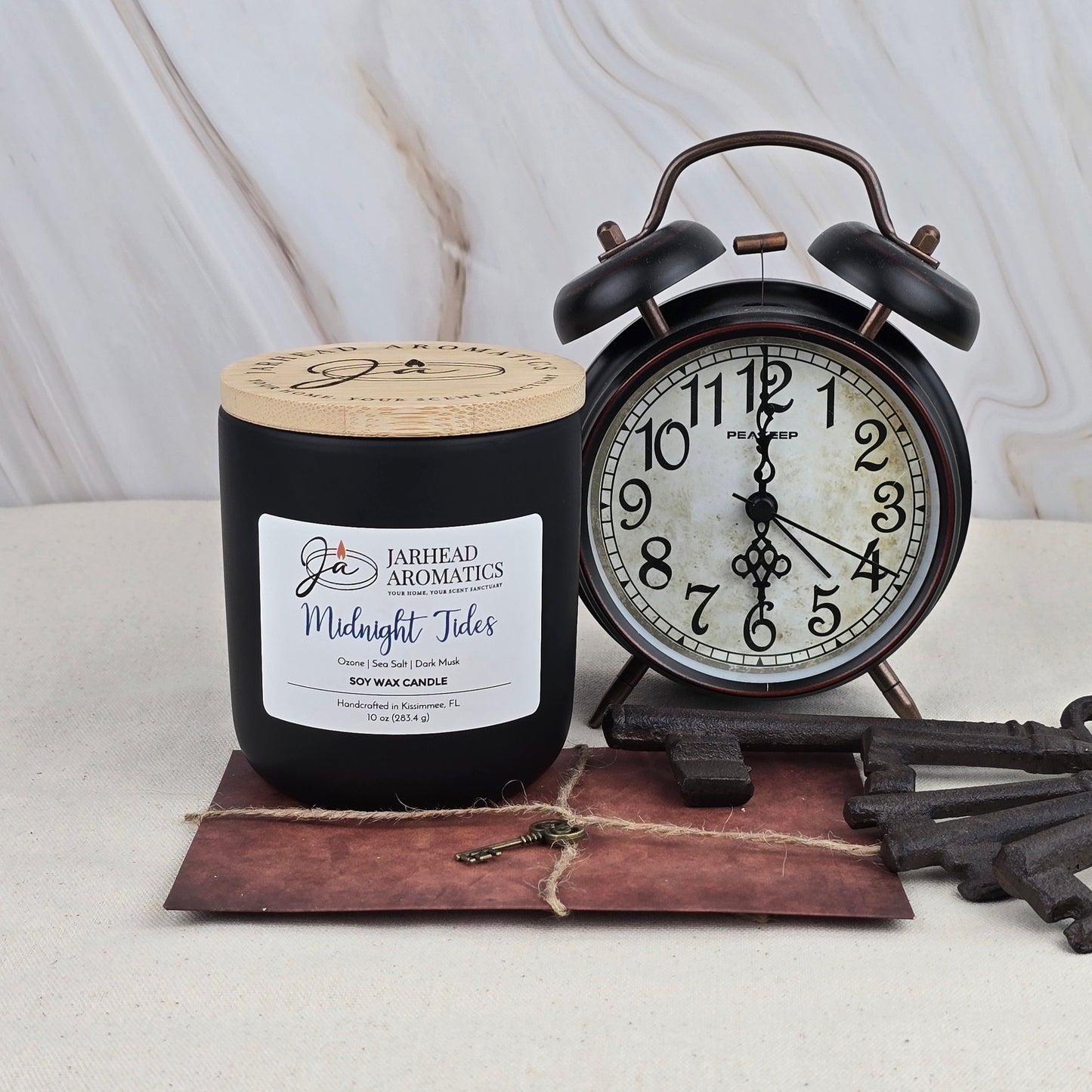 A Jarhead Aromatics Midnight Tides Soy Wax Candle in a black jar, featuring a clean ozonic scent from essential oils, is displayed beside a vintage black alarm clock at 7:05, with twine-tied envelope and antique keys on a beige surface.