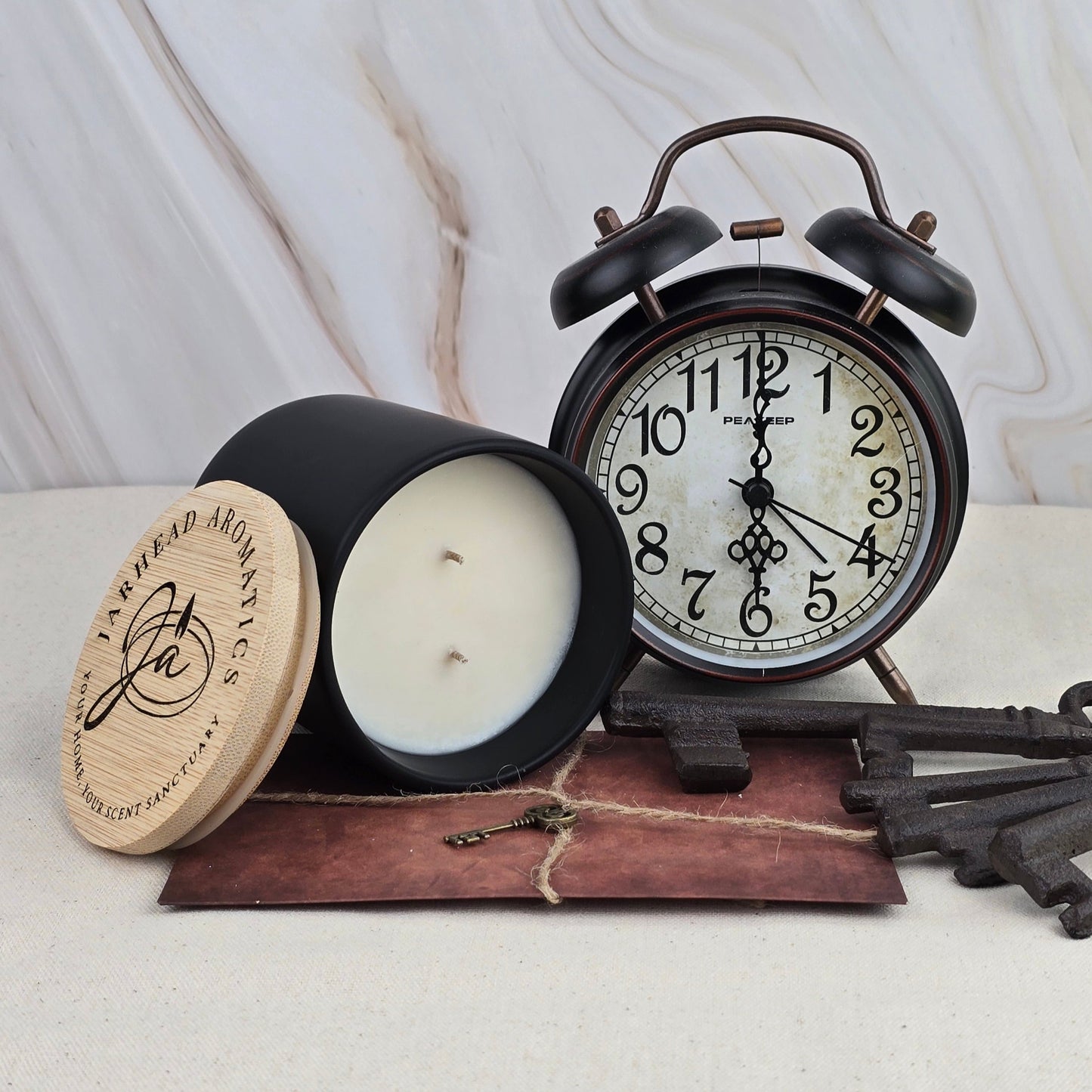 A black vintage alarm clock, the Misty Forest soy wax candle by Jarhead Aromatics, and old-fashioned keys sit on a brown leather pouch against a light marble background.
