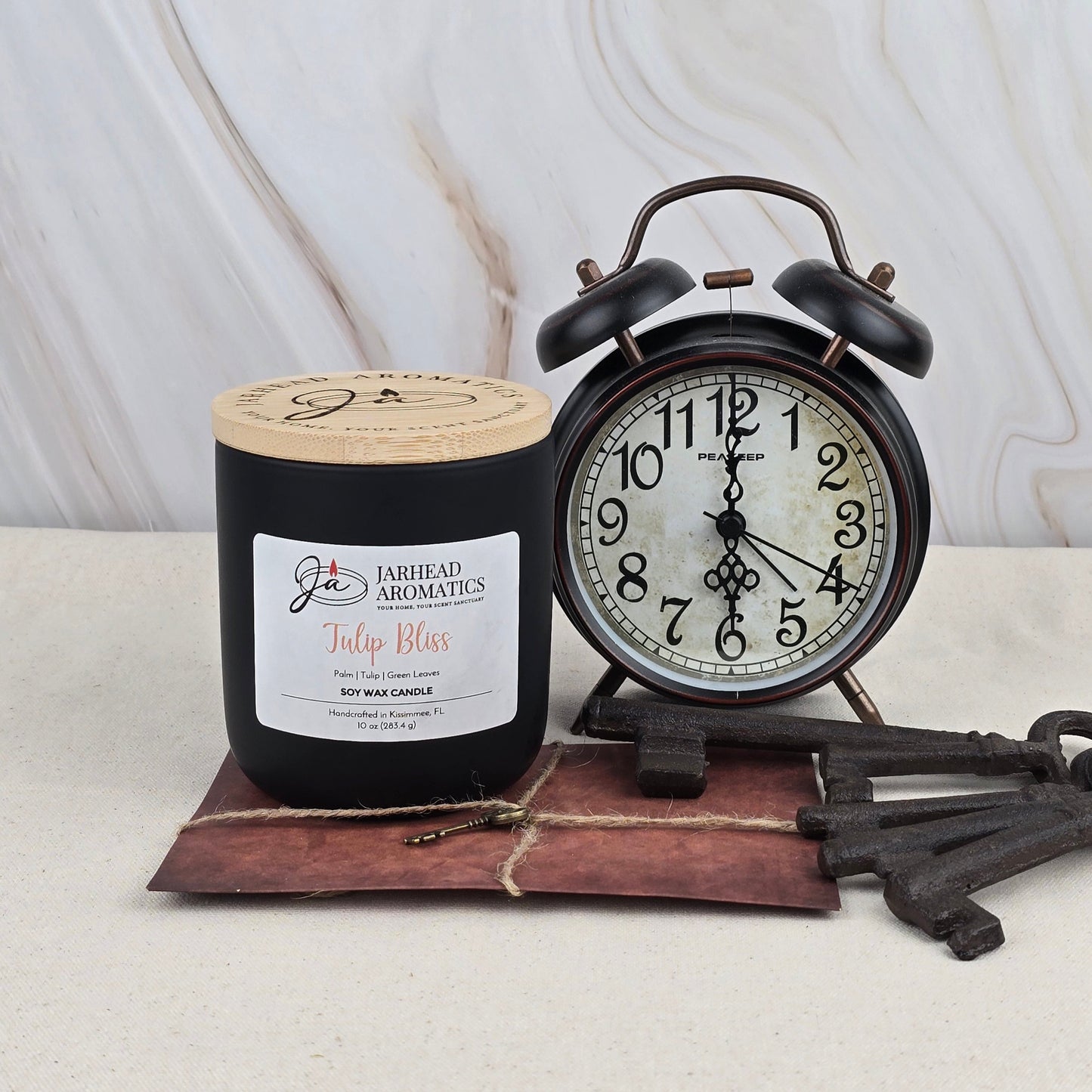 A black Jarhead Aromatics Tulip Bliss - Soy Wax Candle with essential oils sits by a vintage black alarm clock set to 9:10, three old metal keys, and a brown envelope, all atop a light marble surface.