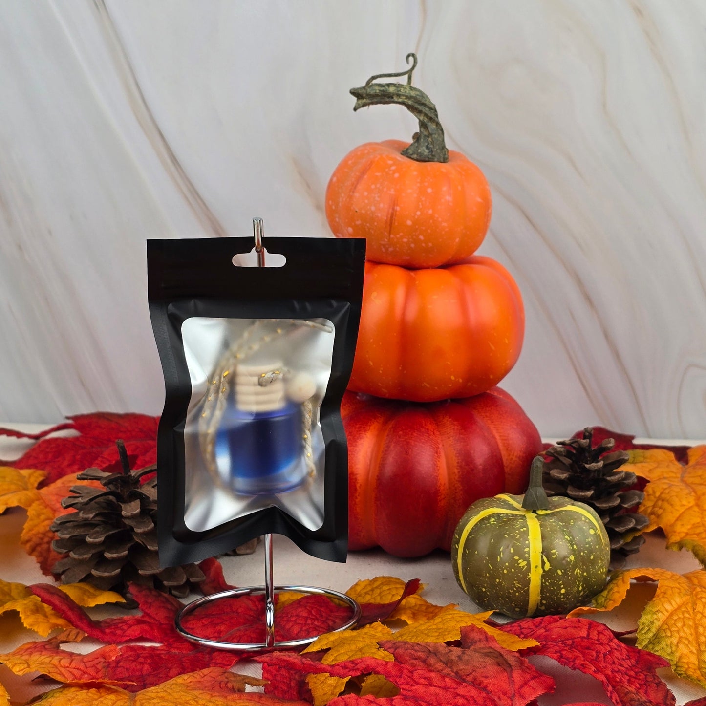 The Cozy Solace Car Diffuser from Jarhead Aromatics’ Fall Collection, in a small blue bottle and black resealable pouch, rests among autumn leaves and pumpkins with an oak moss aroma on a marble-patterned background.