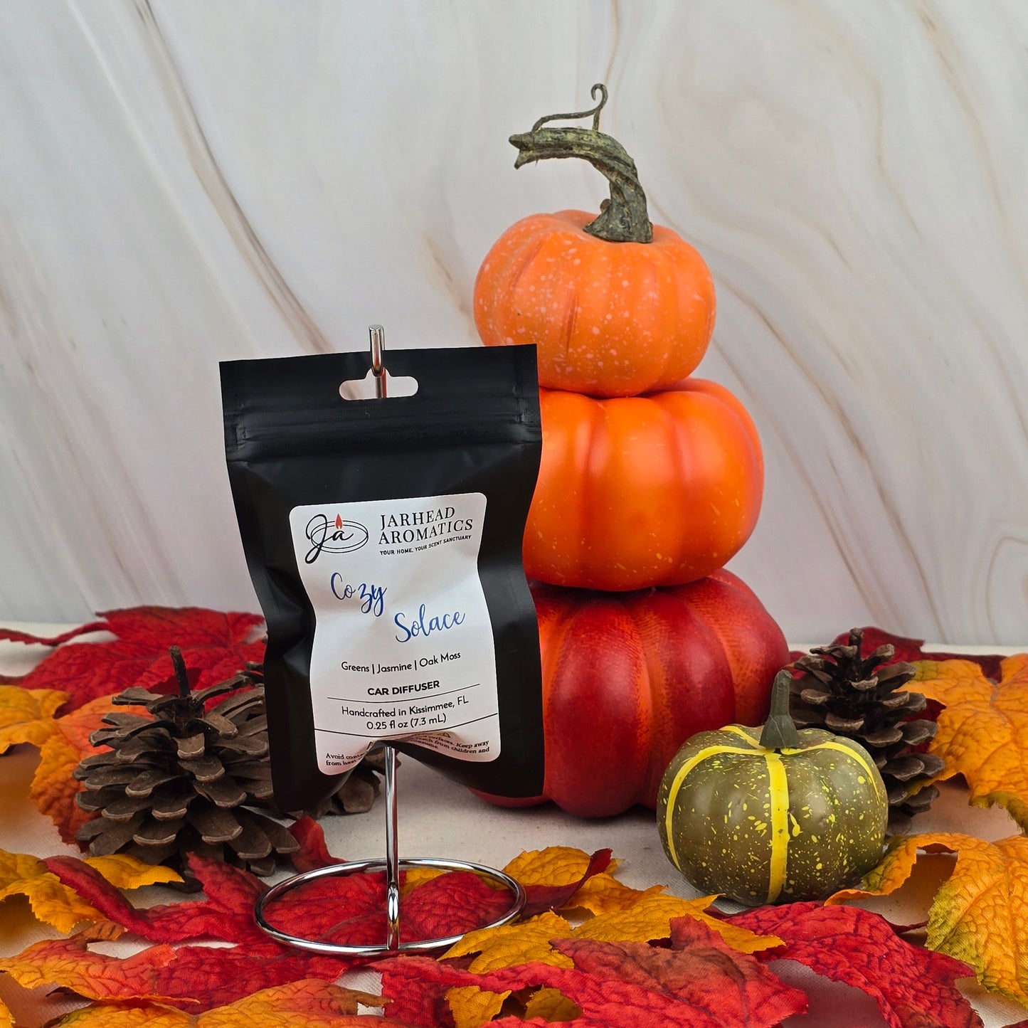 A Cozy Solace Car Diffuser from Jarhead Aromatics Fall Collection is displayed on a holder with autumn leaves, pinecones, green and orange pumpkins, all set against a marble background.