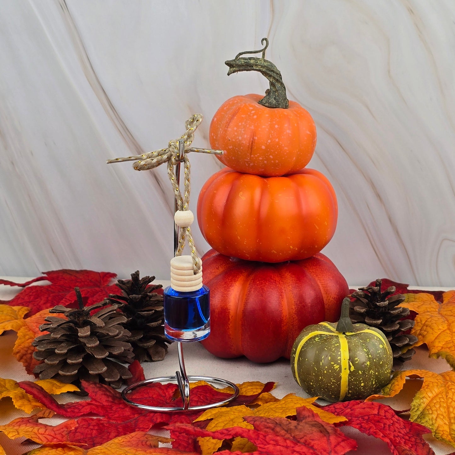 The Cozy Solace Car Diffuser from Jarhead Aromatics Fall Collection features autumn leaves, pinecones, stacked pumpkins, a green pumpkin, and a blue glass ornament with a wooden bead, capturing oak moss freshness over marble accents.