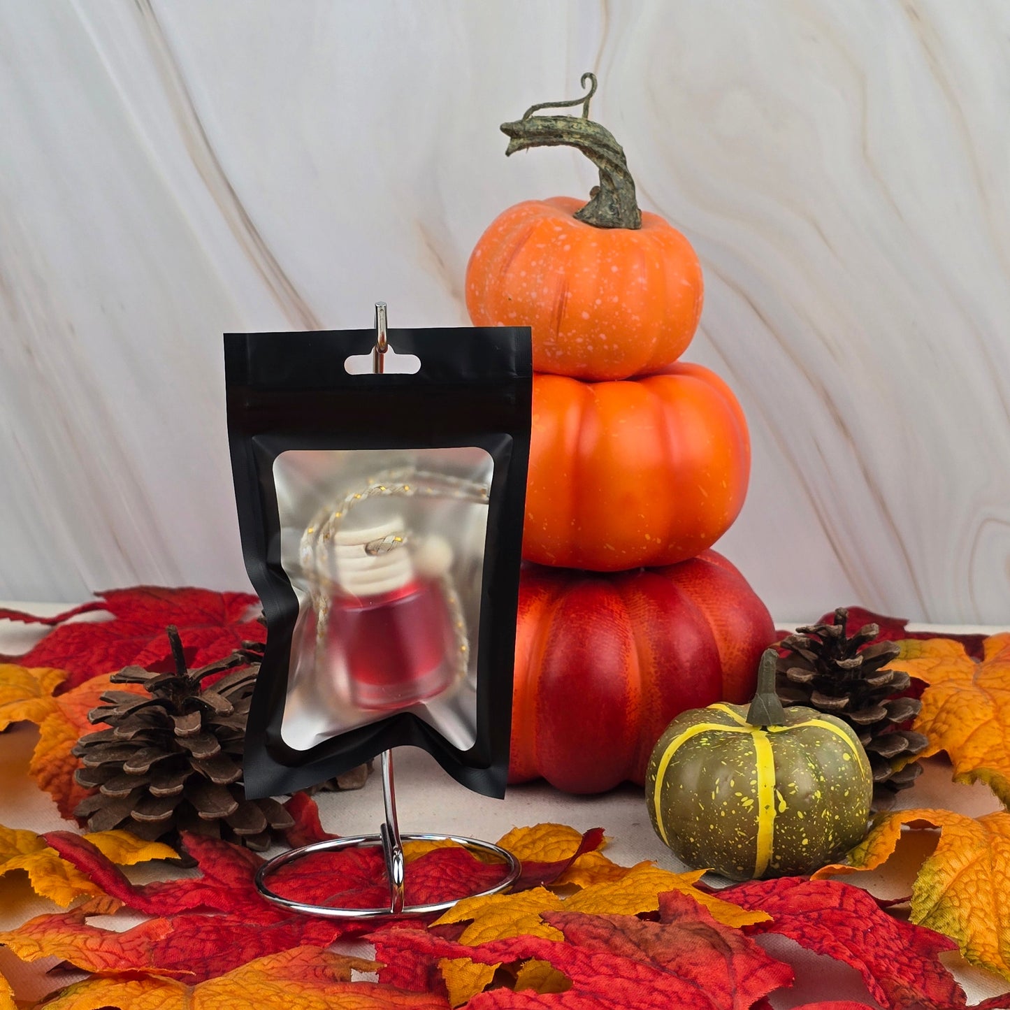 Displayed on a stand is the Jarhead Aromatics Cran-Apple Delight Car Diffuser from the Fall Collection—a small black and clear resealable bag with a red and gold ornament. Stacked pumpkins, pinecones, and autumn leaves create a festive background.