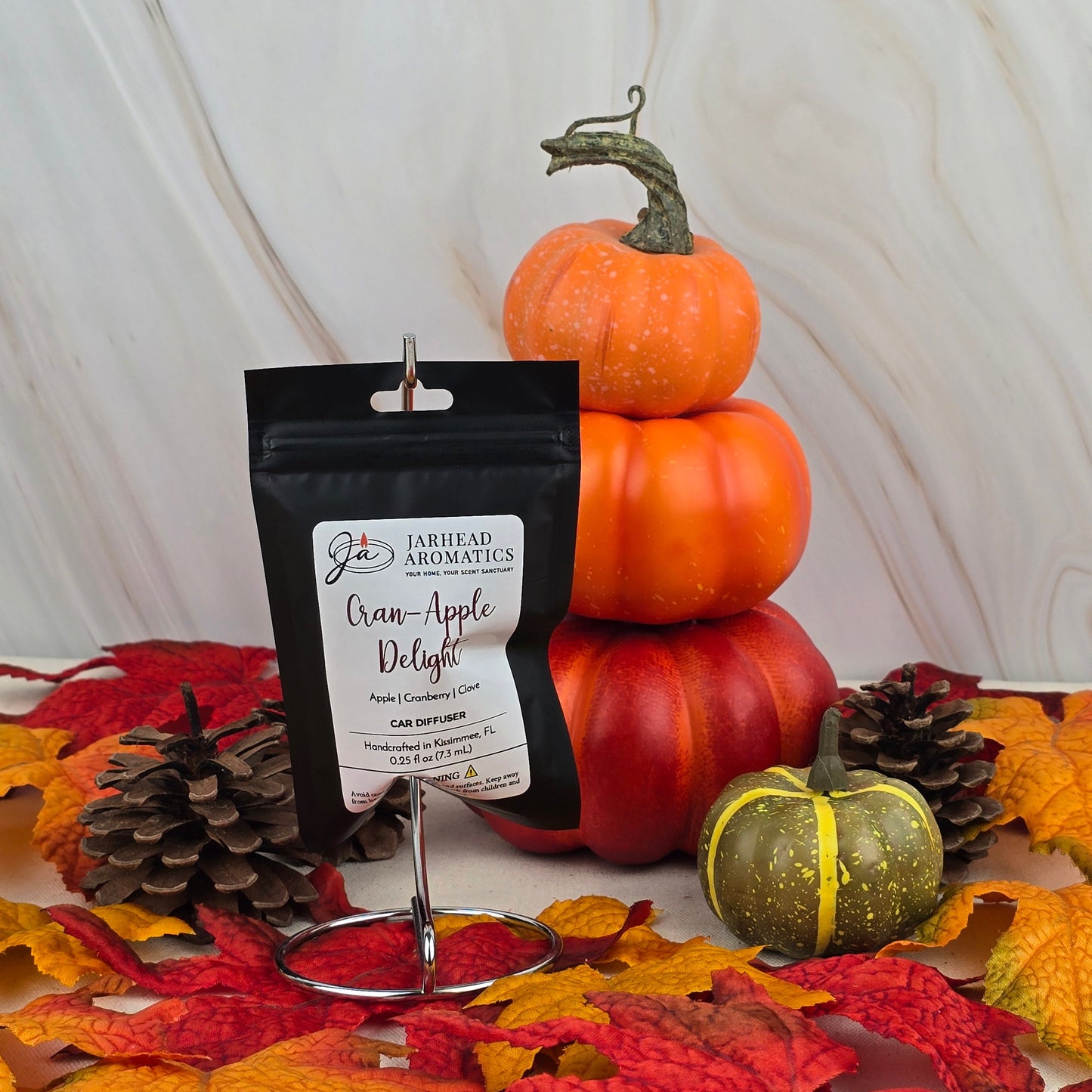A Jarhead Aromatics Cran-Apple Delight Car Diffuser from the Fall Collection is displayed among small pumpkins, pinecones, and autumn leaves, creating a festive seasonal scene.