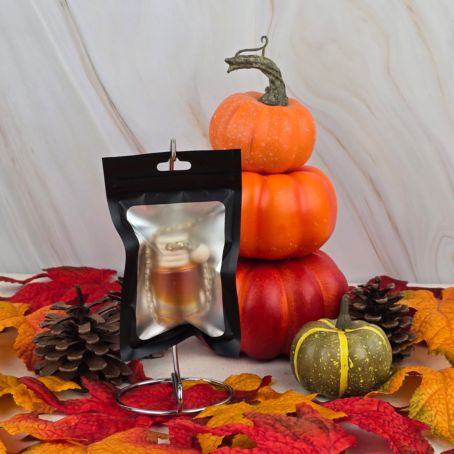 A small black pouch with a clear window displays the Harvest Cream Car Diffuser by Jarhead Aromatics. Set on a metal holder with pumpkins, pinecones, and autumn leaves, it brings cozy fall fragrance to your drive.