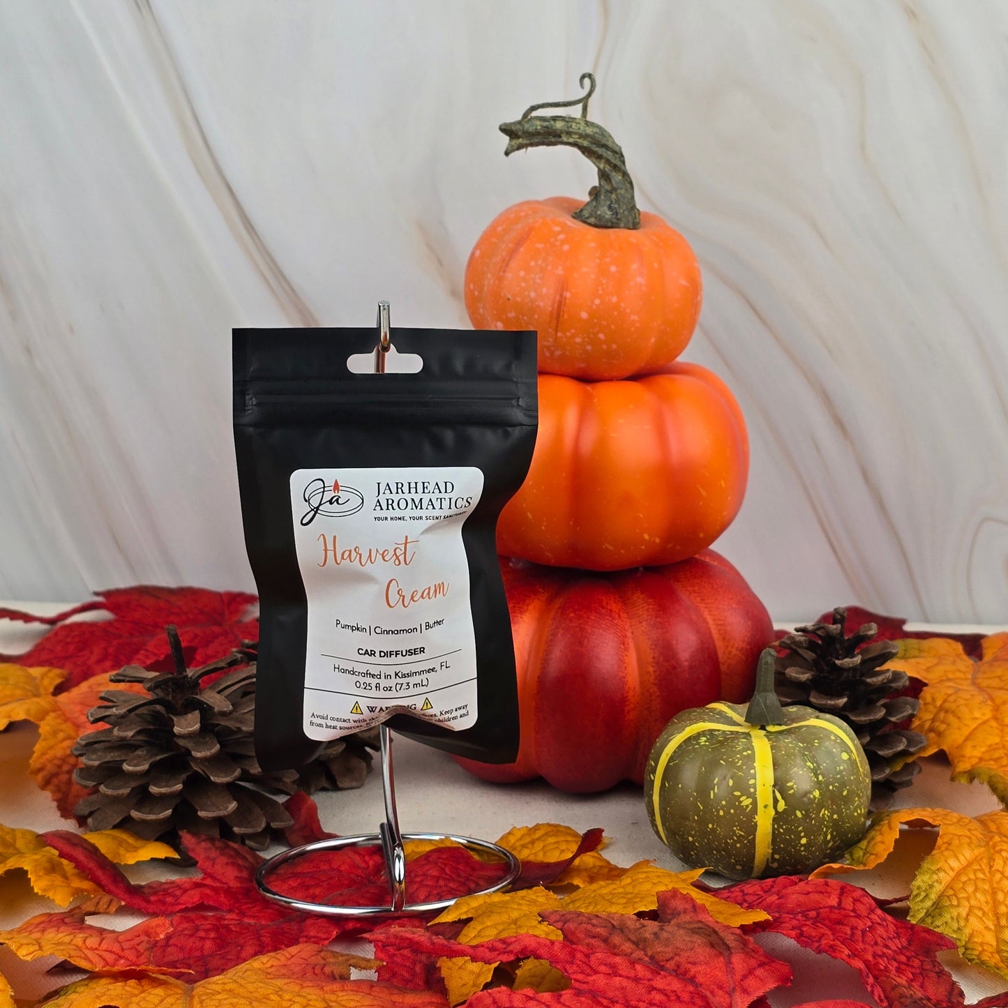 The Jarhead Aromatics Harvest Cream Car Diffuser from the Fall Collection rests on a metal stand, encircled by pinecones, autumn leaves, and pumpkins against a marbled background.