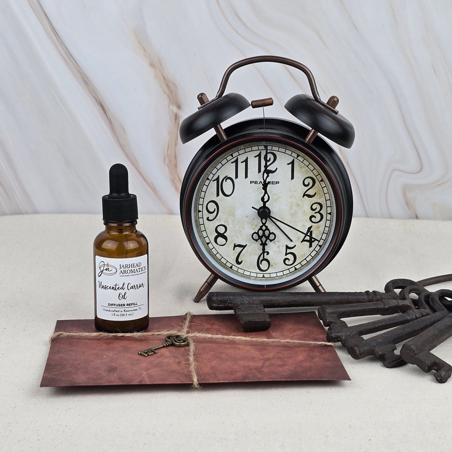 A brown glass bottle of Jarhead Aromatics Unscented Carrier Oil - Car Diffuser Refill is arranged with a vintage alarm clock, old-fashioned keys, and a tied brown envelope on a light surface against a marble-patterned background.