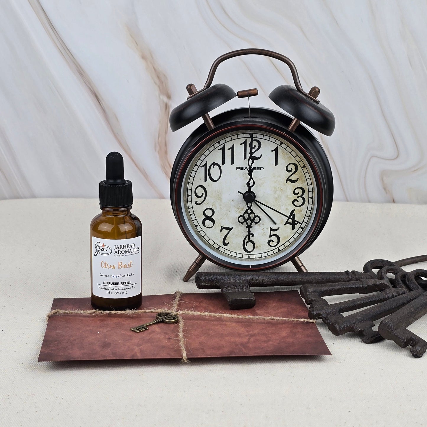 A Citrus Burst Car Diffuser Refill by Jarhead Aromatics rests on a light surface with a marble-patterned background, accompanied by a vintage alarm clock, brown string-tied envelope, and old metal keys.