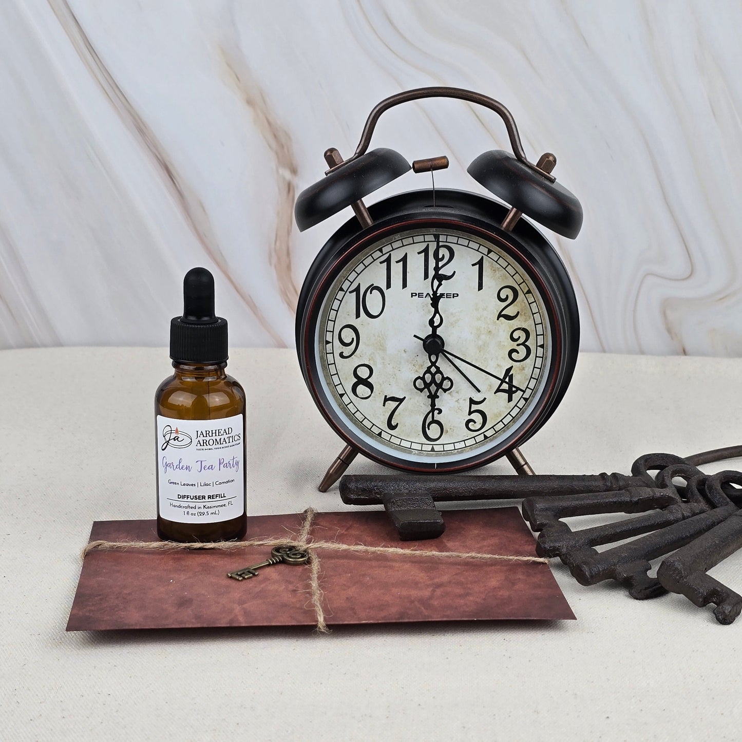 A Garden Tea Party Car Diffuser Refill by Jarhead Aromatics sits on a light surface with a marble-patterned background, surrounded by an alarm clock, amber dropper bottle, old keys, and a tied brown envelope.