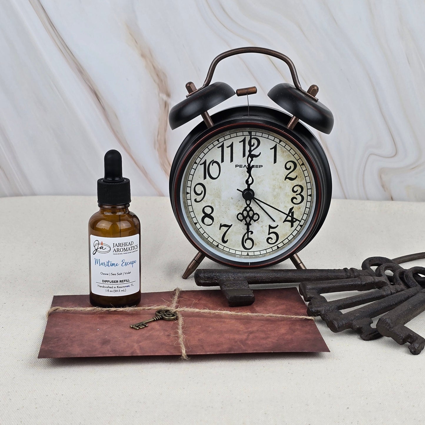 A vintage alarm clock, old-fashioned keys, and a brown envelope surround Jarhead Aromatics Maritime Escape - Car Diffuser Refill, a dropper bottle with natural essential oils, displayed on a light surface against a marbled backdrop.