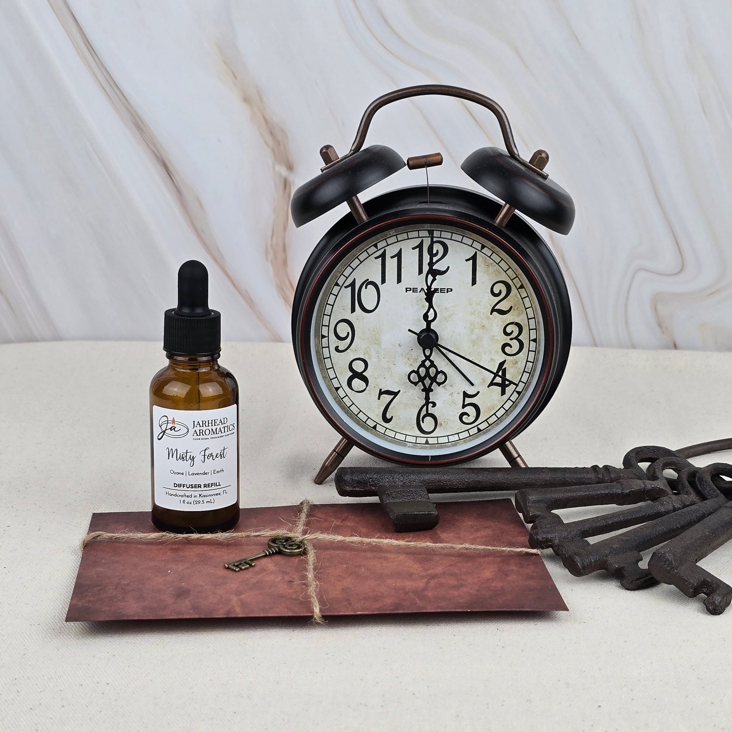 A Misty Forest - Car Diffuser Refill by Jarhead Aromatics, a vintage alarm clock, an envelope tied with string, and large old-fashioned keys are arranged on a light surface with a marble background.