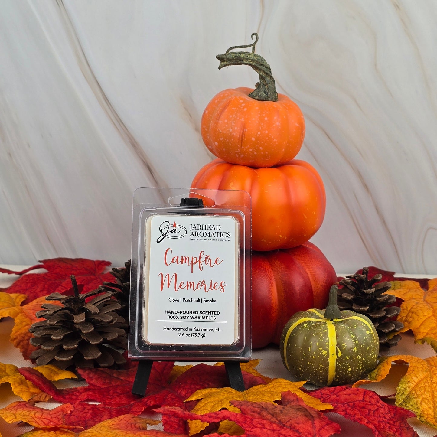 A Jarhead Aromatics Campfire Memories - Soy Wax Melt with a smoky scent is displayed on a small easel, surrounded by pinecones, faux pumpkins, and autumn leaves against a light background.