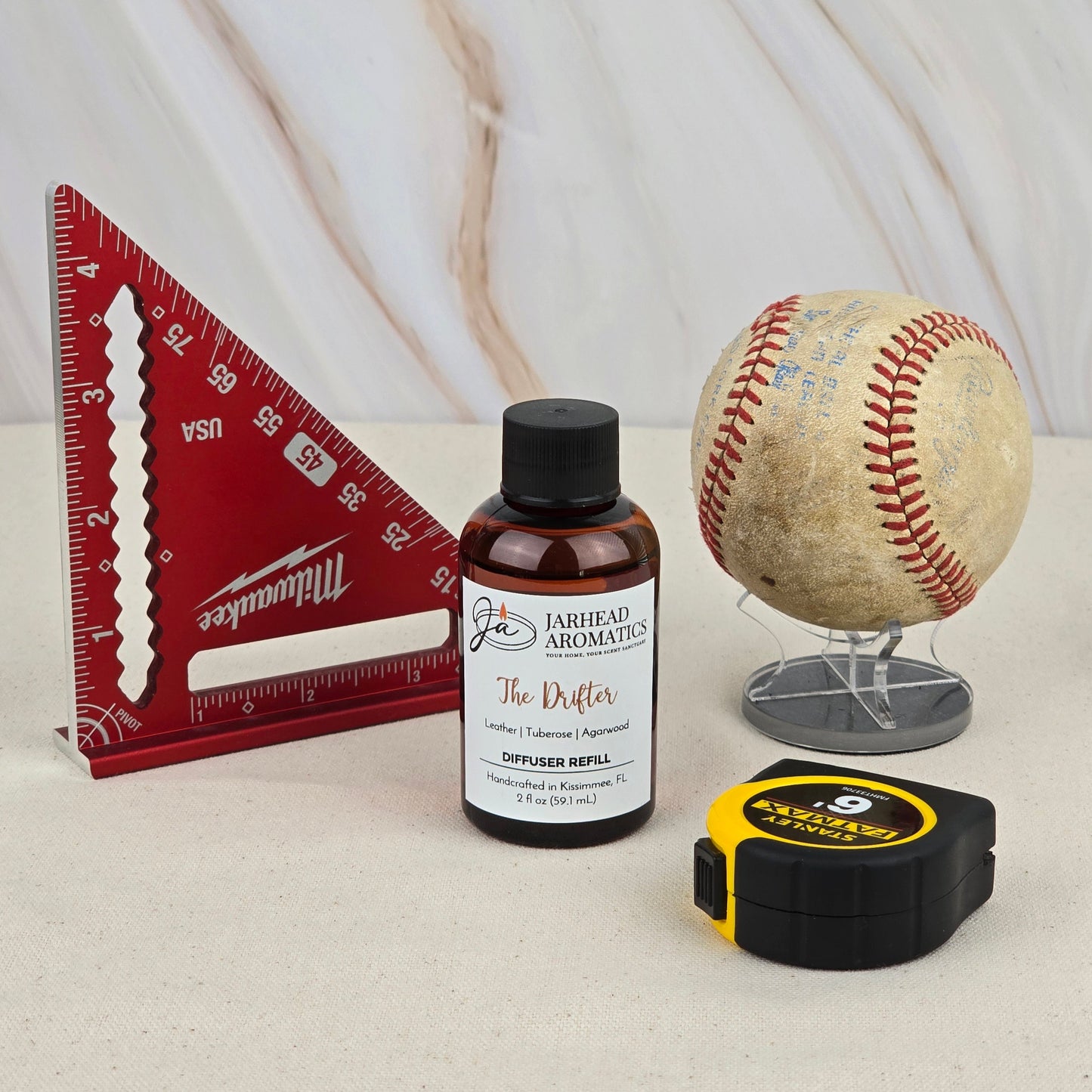 A red triangular ruler, The Drifter Diffuser Refill by Jarhead Aromatics, a worn baseball, and a yellow-black tape measure suggest the wild frontier, set on a beige surface with a marbled background.