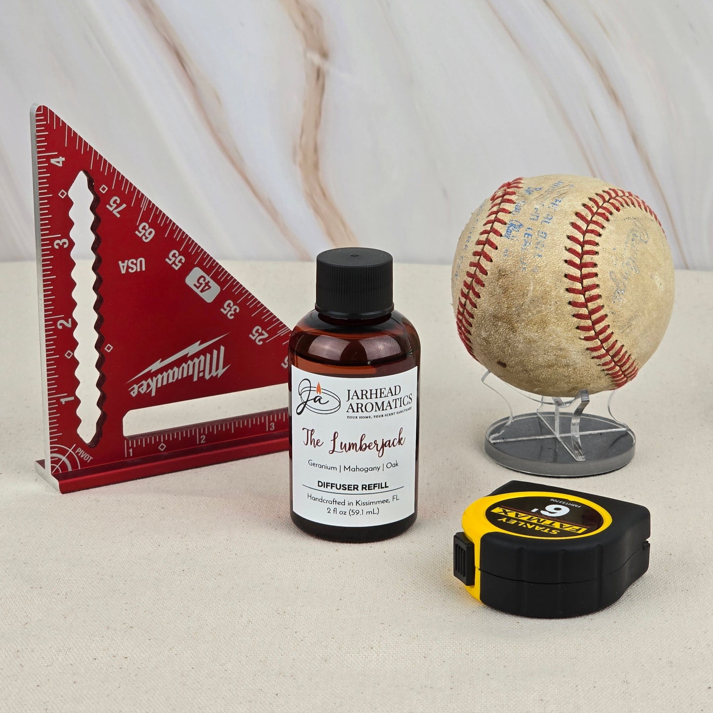 A red triangular ruler, an old baseball, a yellow-black tape measure, and a Jarhead Aromatics The Lumberjack - Diffuser Refill are arranged on beige with a marble backdrop, creating an outdoors-inspired scene.