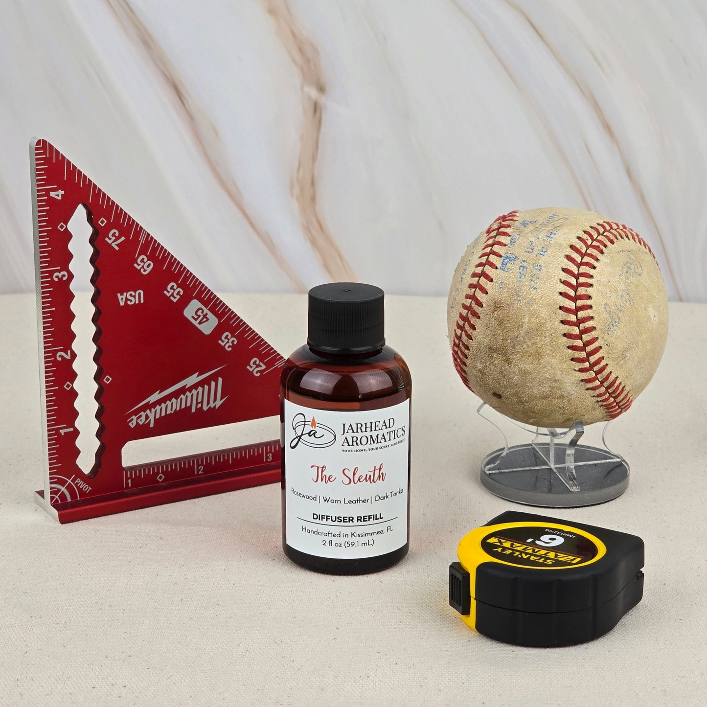 A red triangle ruler, a worn baseball on a stand, a yellow tape measure, and Jarhead Aromatics’ The Sleuth - Diffuser Refill—radiating a warm, woody scent—are arranged on a light surface with a marble backdrop.