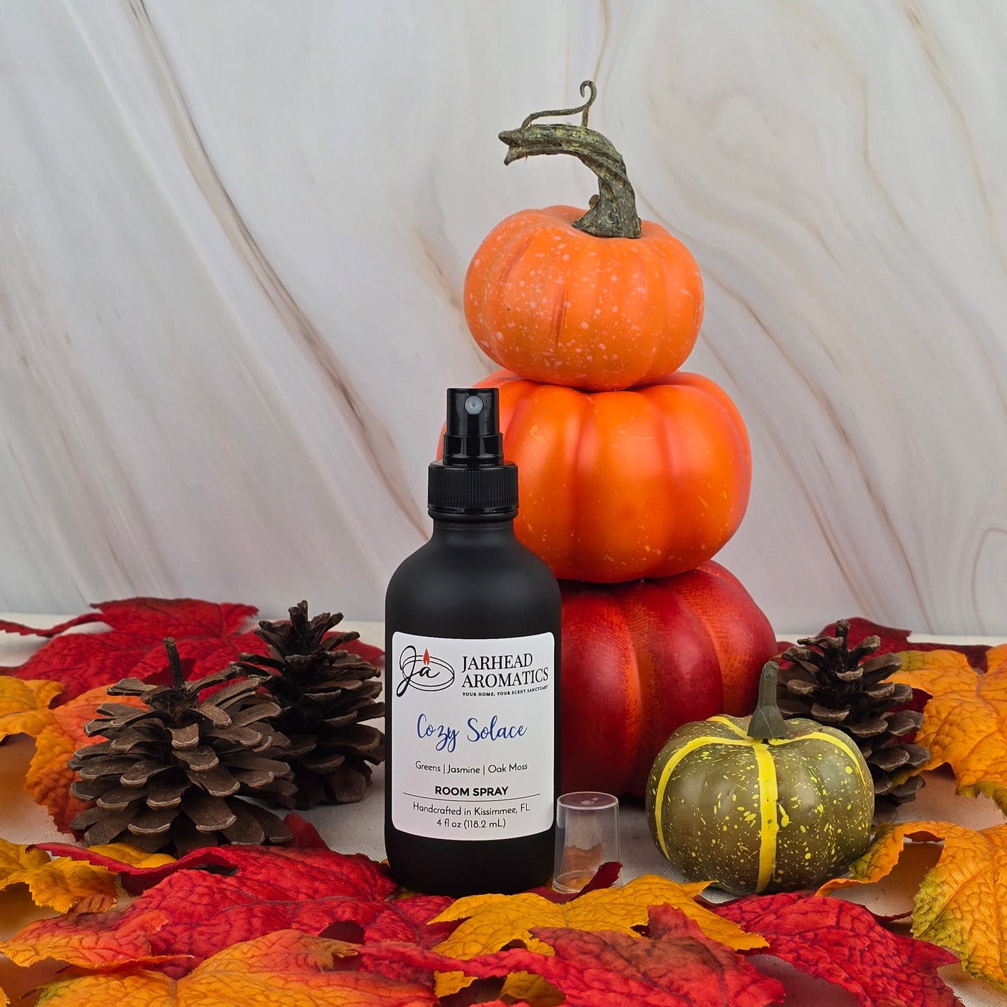A black bottle of Jarhead Aromatics Cozy Solace Room Spray—Fall Collection is surrounded by autumn leaves, pinecones, and stacked pumpkins on a marbled background, evoking scents of jasmine and fresh greens.