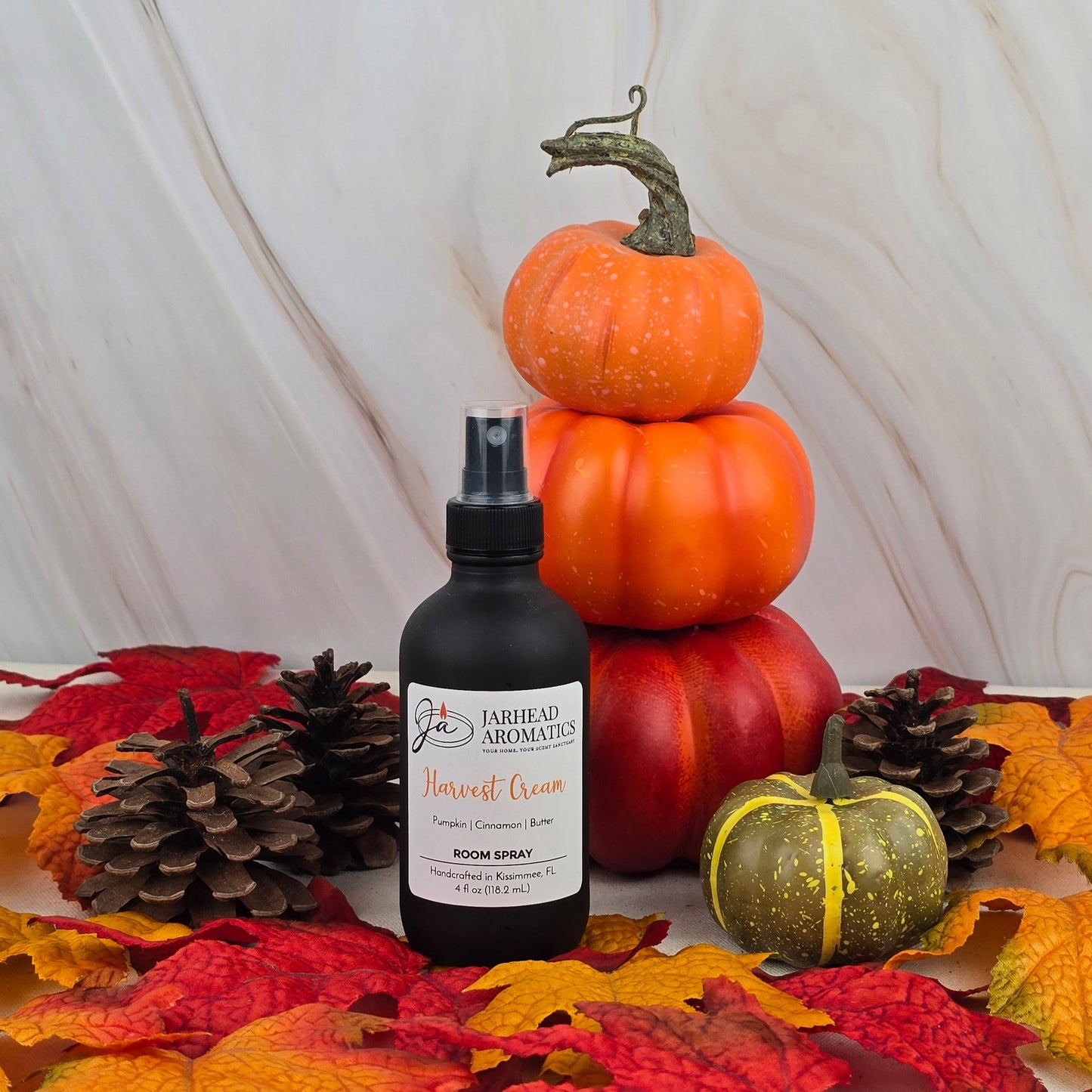 A bottle of Jarhead Aromatics Harvest Cream Room Spray from the Fall Collection sits among pinecones, faux autumn leaves, and stacked pumpkins, adding a cozy fall scent to the marble-backed festive display.