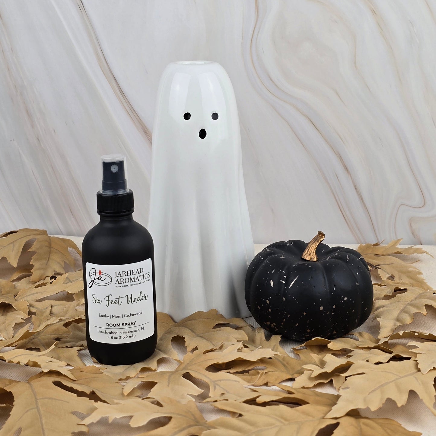 A ceramic ghost, a black pumpkin, and a bottle of Six Feet Under Room Spray by Jarhead Aromatics are arranged on dried oak leaves in front of a marbled background.