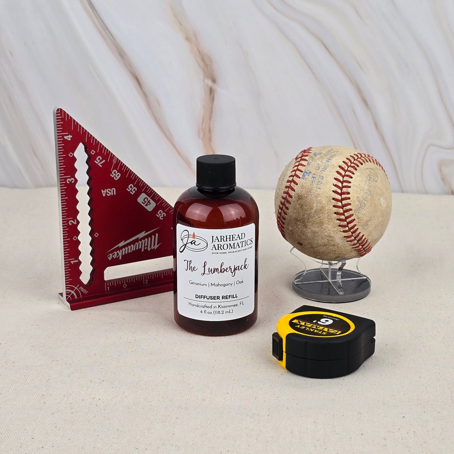 A red triangular ruler, Jarhead Aromatics’ The Lumberjack - Diffuser Refill, a worn baseball on a stand, and a black and yellow tape measure are displayed on a beige surface against a marble-patterned background.