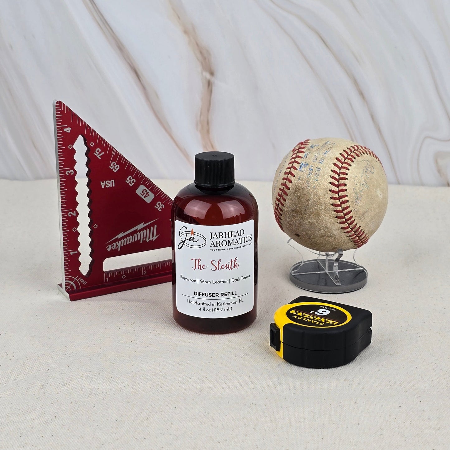 A bottle of Jarhead Aromatics The Sleuth Diffuser Refill, a red triangular ruler, a baseball on a stand, and a yellow tape measure are arranged on a light surface with a marble-patterned background.