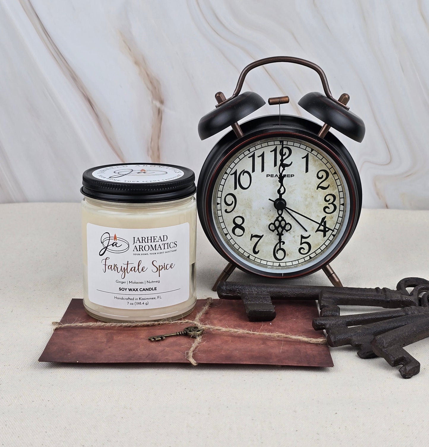 A black vintage alarm clock, a Jarhead Aromatics Fairytale Spice Soy Wax Candle with gingerbread-like notes, an old envelope tied with twine, and two ornate metal keys rest on a light marbled background.