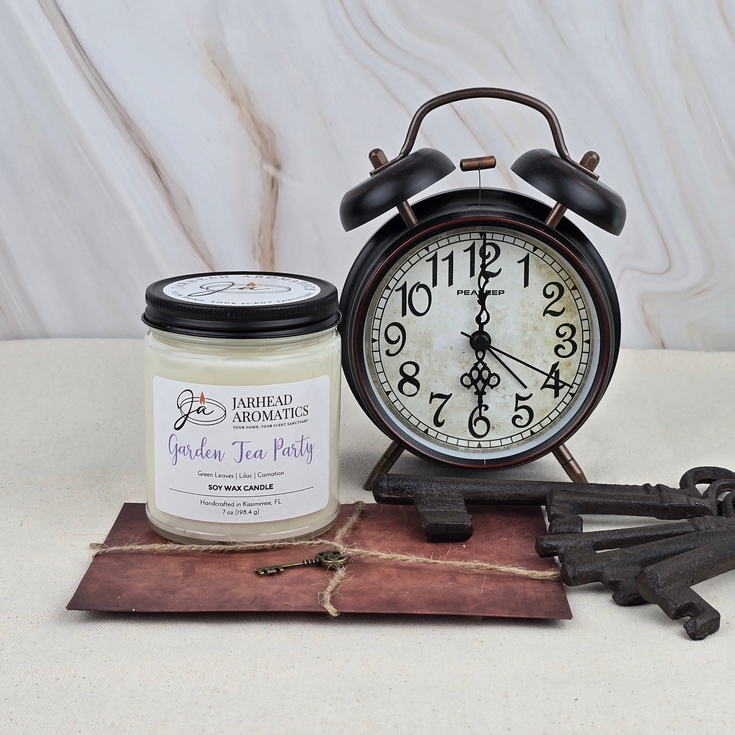 A Jarhead Aromatics Garden Tea Party Soy Wax Candle with citrus scent, a vintage black alarm clock at 7:00, a twine-tied brown envelope, and three old keys are arranged on marble against a light background.