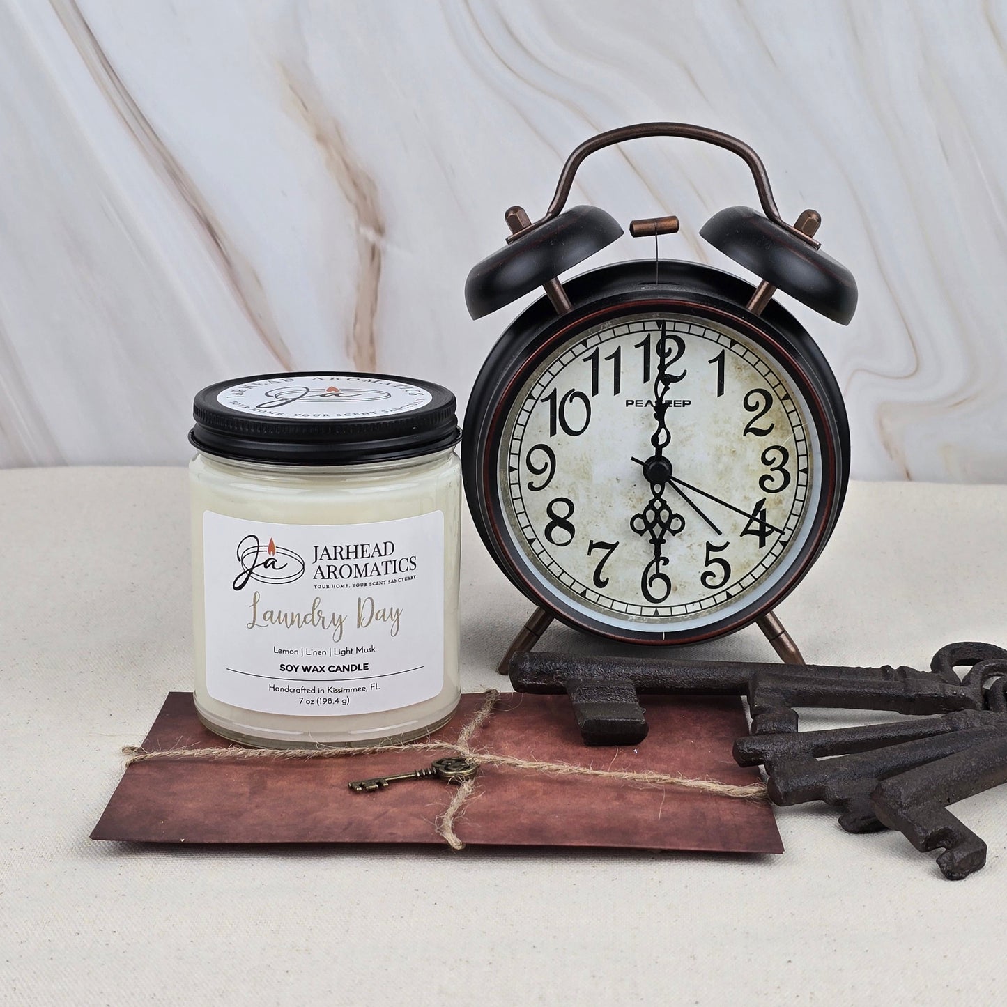 A vintage black alarm clock displays 8:02 beside a Jarhead Aromatics Laundry Day - Soy Wax Candle in a glass jar, a twine-tied brown envelope, and three antique-style keys on a marble surface.