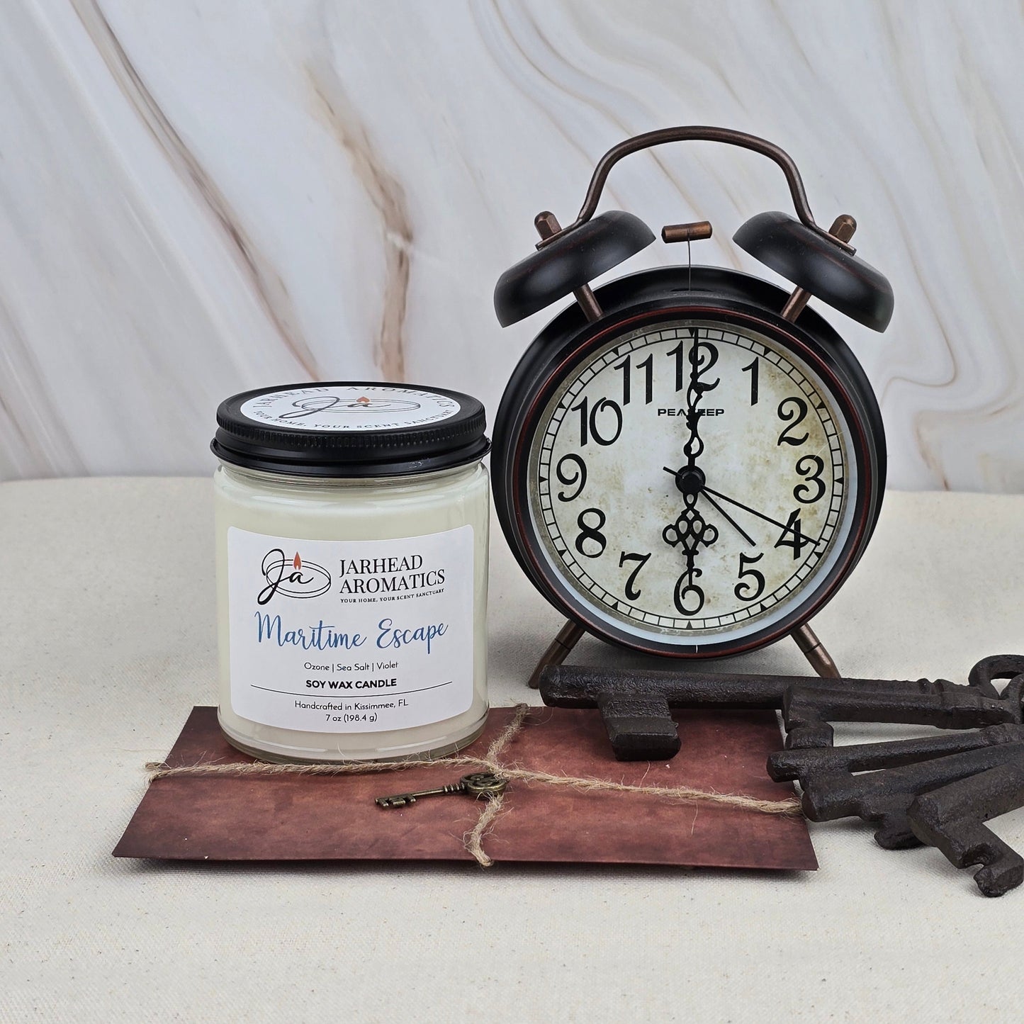 The Maritime Escape - Soy Wax Candle by Jarhead Aromatics sits beside a vintage black alarm clock on a rustic brown tray adorned with decorative old keys, all set against a light marble background.