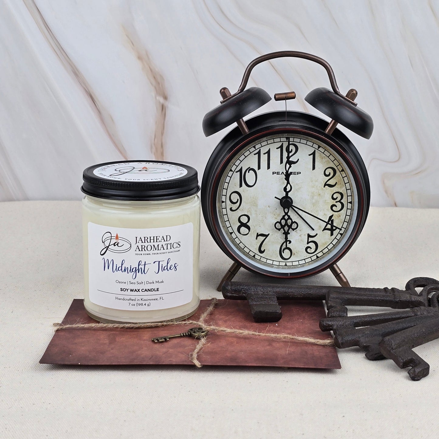 A black vintage alarm clock at 10:10 rests beside Jarhead Aromatics’ Midnight Tides Soy Wax Candle, its fresh, essential oil-like scent emanating from a wax-sealed envelope and two large antique keys on a light surface.