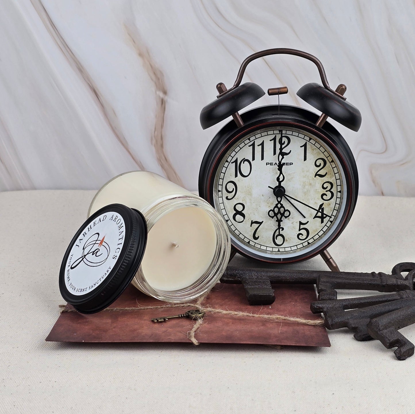 A vintage black alarm clock sits beside Jarhead Aromatics Misty Forest soy wax candle, infused with earthy, woodsy scents in a labeled glass jar, placed on a leather-bound book with antique keys against a light marble background.