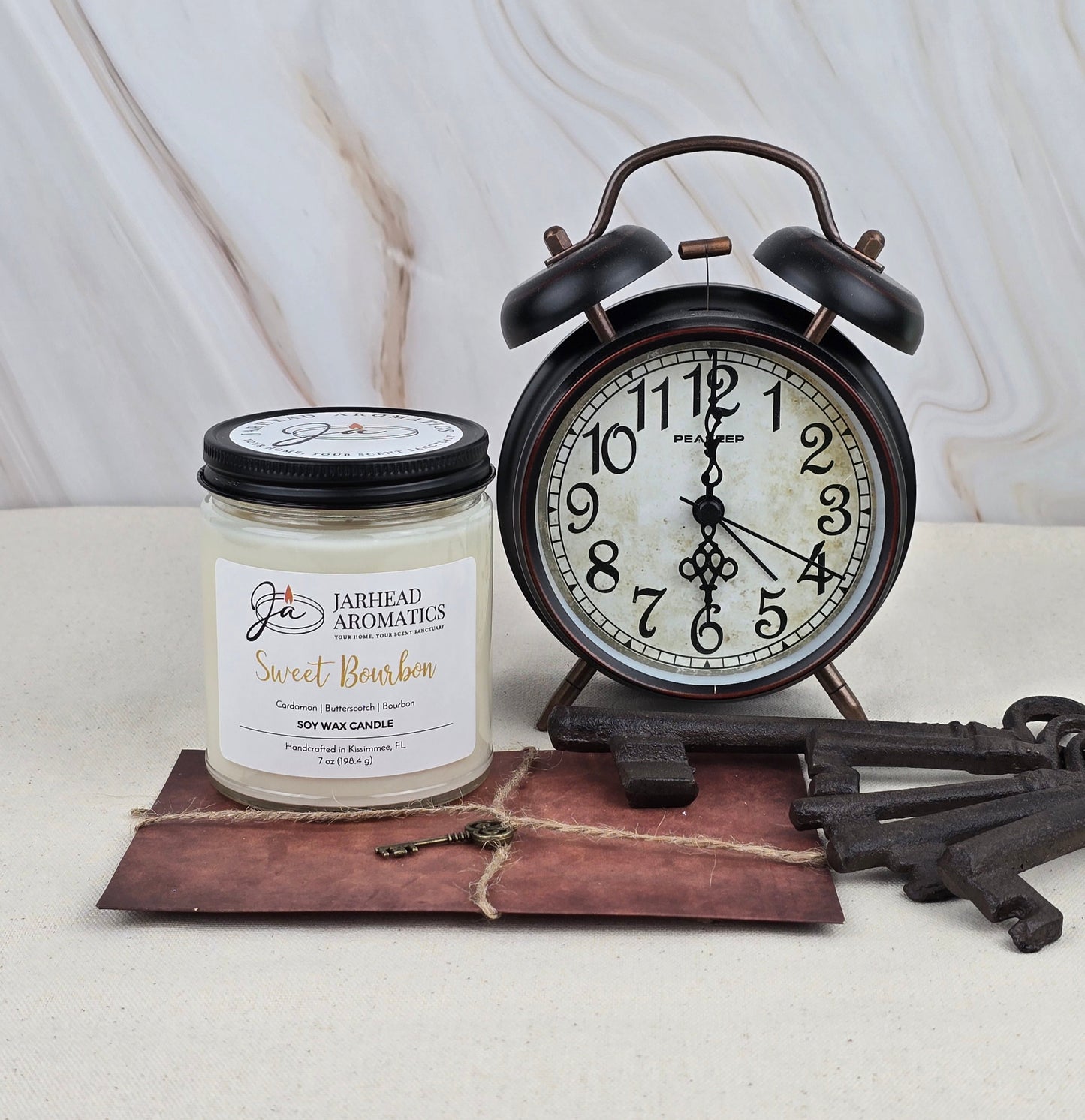 A black vintage alarm clock, a Jarhead Aromatics Sweet Bourbon - Soy Wax Candle, a twine-tied brown envelope, and three antique keys rest on a light surface against a neutral marbled background.