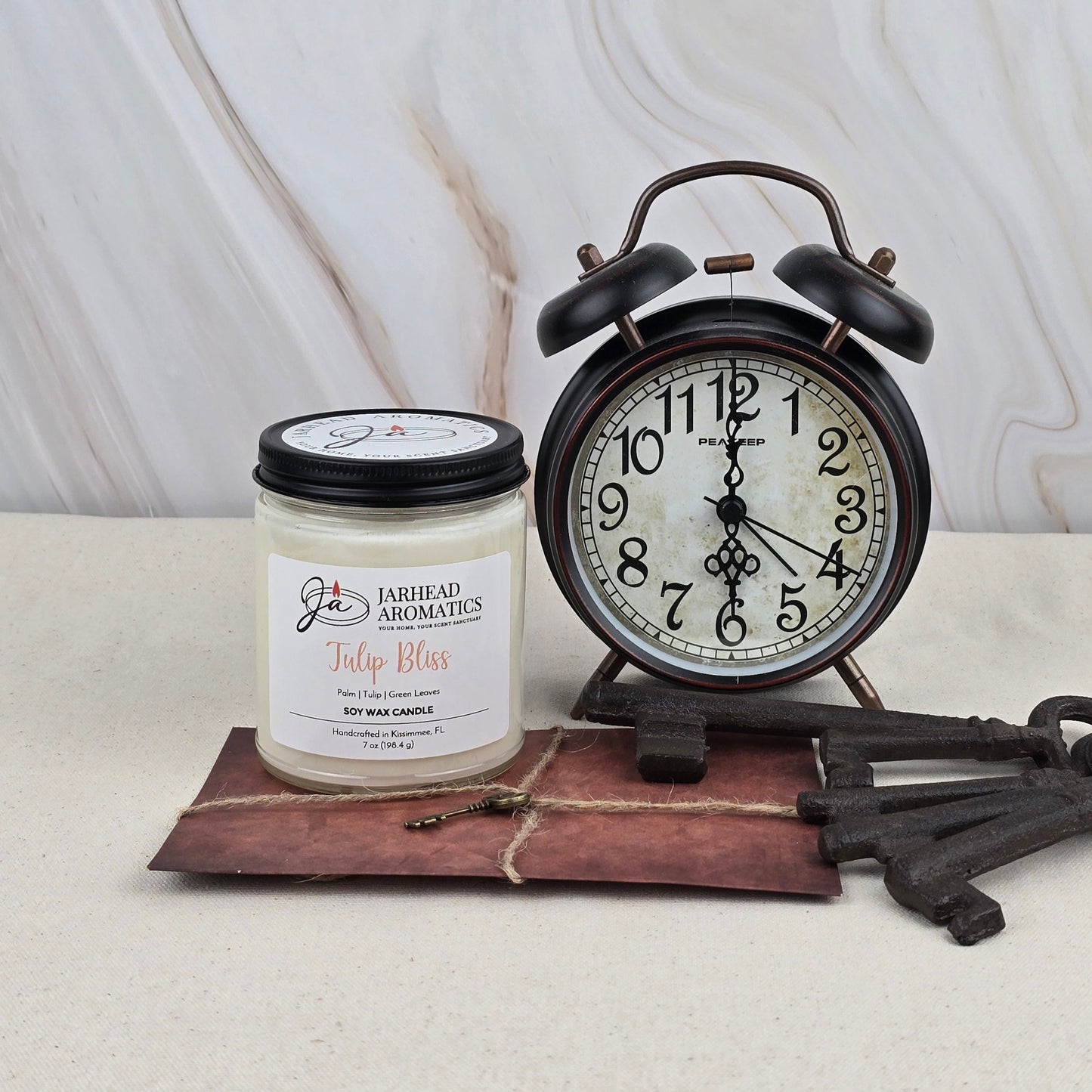 A black vintage alarm clock sits beside a Jarhead Aromatics Tulip Bliss Soy Wax Candle on a brown leather-bound journal with a key, set against a light marble background.
