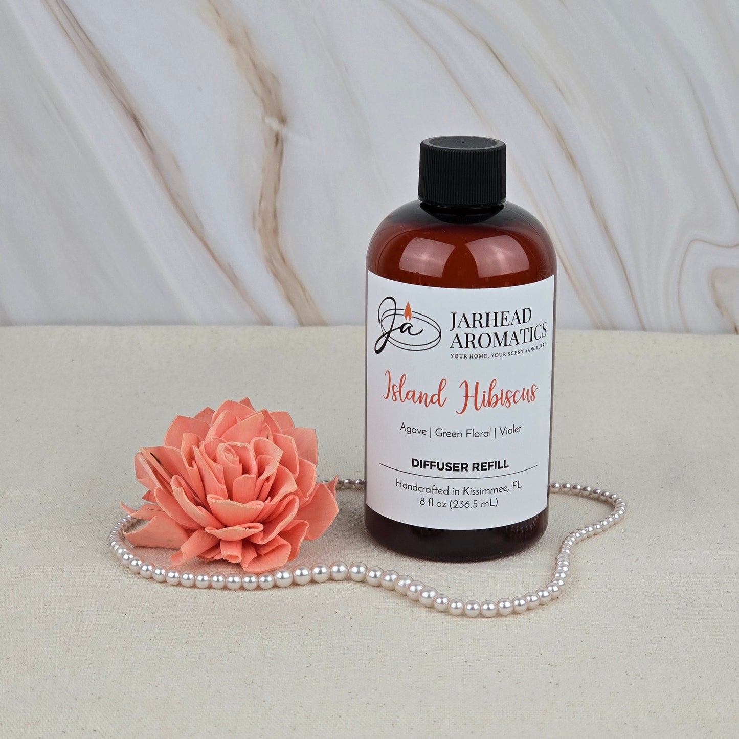 A bottle of Jarhead Aromatics Island Hibiscus - Diffuser Refill sits beside a coral artificial flower and pearls on a beige surface with a marble backdrop, evoking the vibe of a tropical paradise.