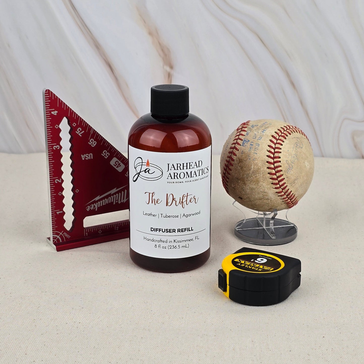 A brown bottle labeled Jarhead Aromatics The Drifter - Diffuser Refill, featuring rugged leather notes, sits on a light surface beside a red triangular ruler, a worn baseball on a stand, and a black-and-yellow tape measure. Marble background.