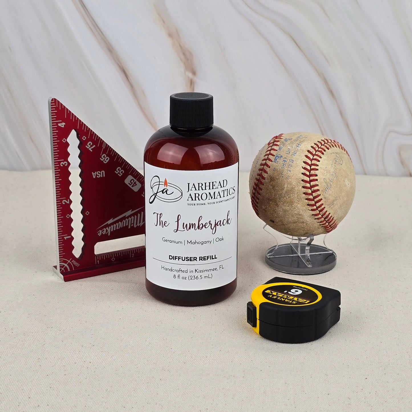 A bottle of Jarhead Aromatics The Lumberjack - Diffuser Refill rests on a light surface, releasing a woodsy scent beside a red triangular ruler, a vintage baseball on a stand, and a yellow-and-black tape measure.