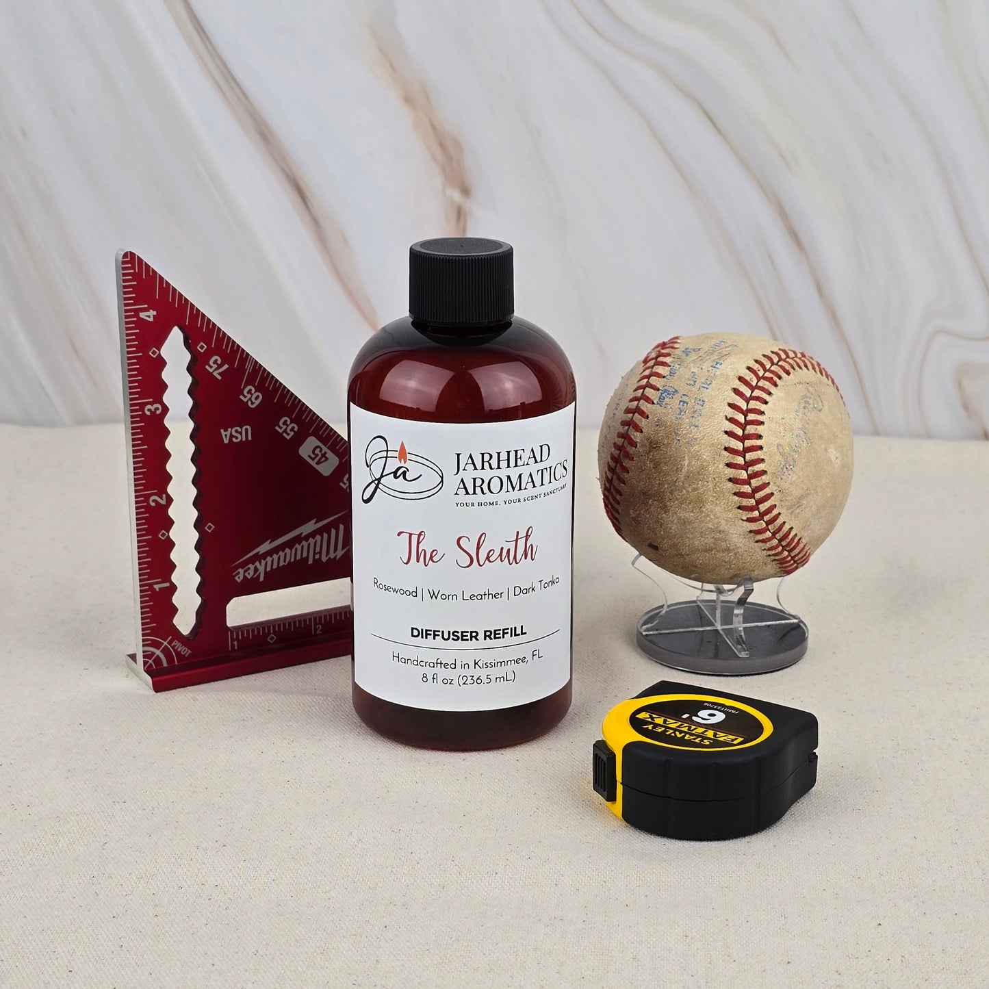 A bottle of The Sleuth - Diffuser Refill by Jarhead Aromatics sits on a surface beside a red triangular ruler, a worn baseball on a stand, and a black and yellow tape measure, all set against a light marble background.