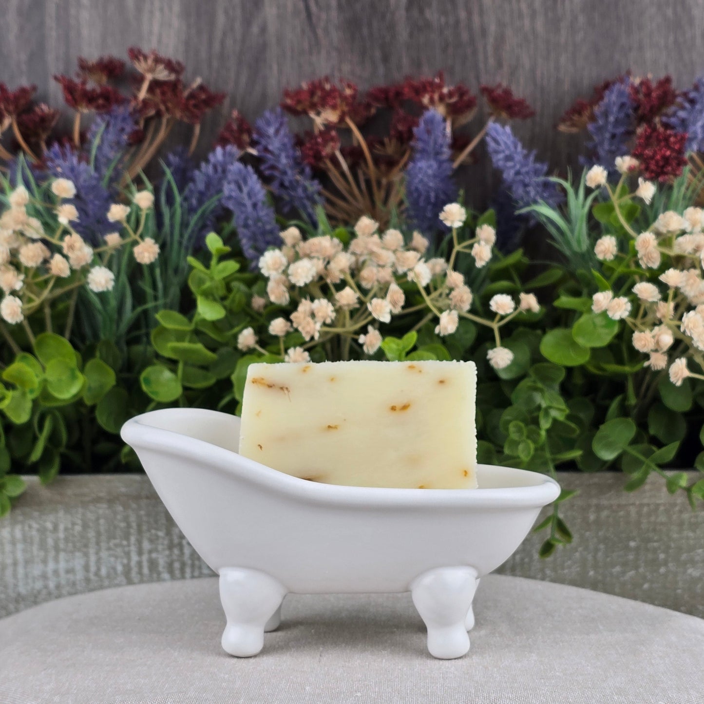 A bar of Jarhead Aromatics Aloe + Calendula Handmade Soap โ All Natural rests in a small, white, bathtub-shaped soap dish with artificial flowers and greenery in the background.