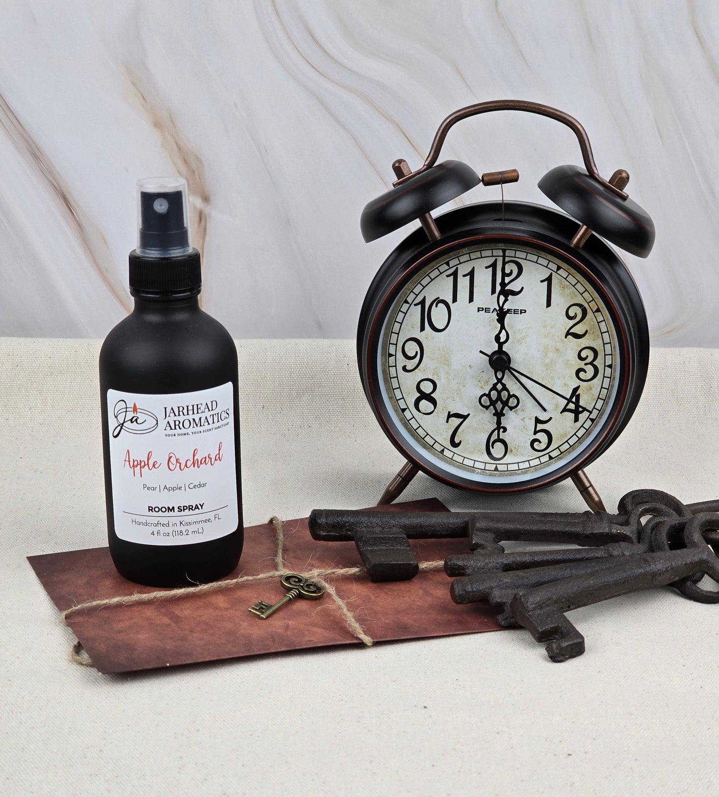 A vintage black alarm clock rests beside Jarhead Aromatics Apple Orchard room spray, infused with fresh apple fragrance, on a brown leather-like mat. Three antique keys and a small brass key complement them against a light marble background.
