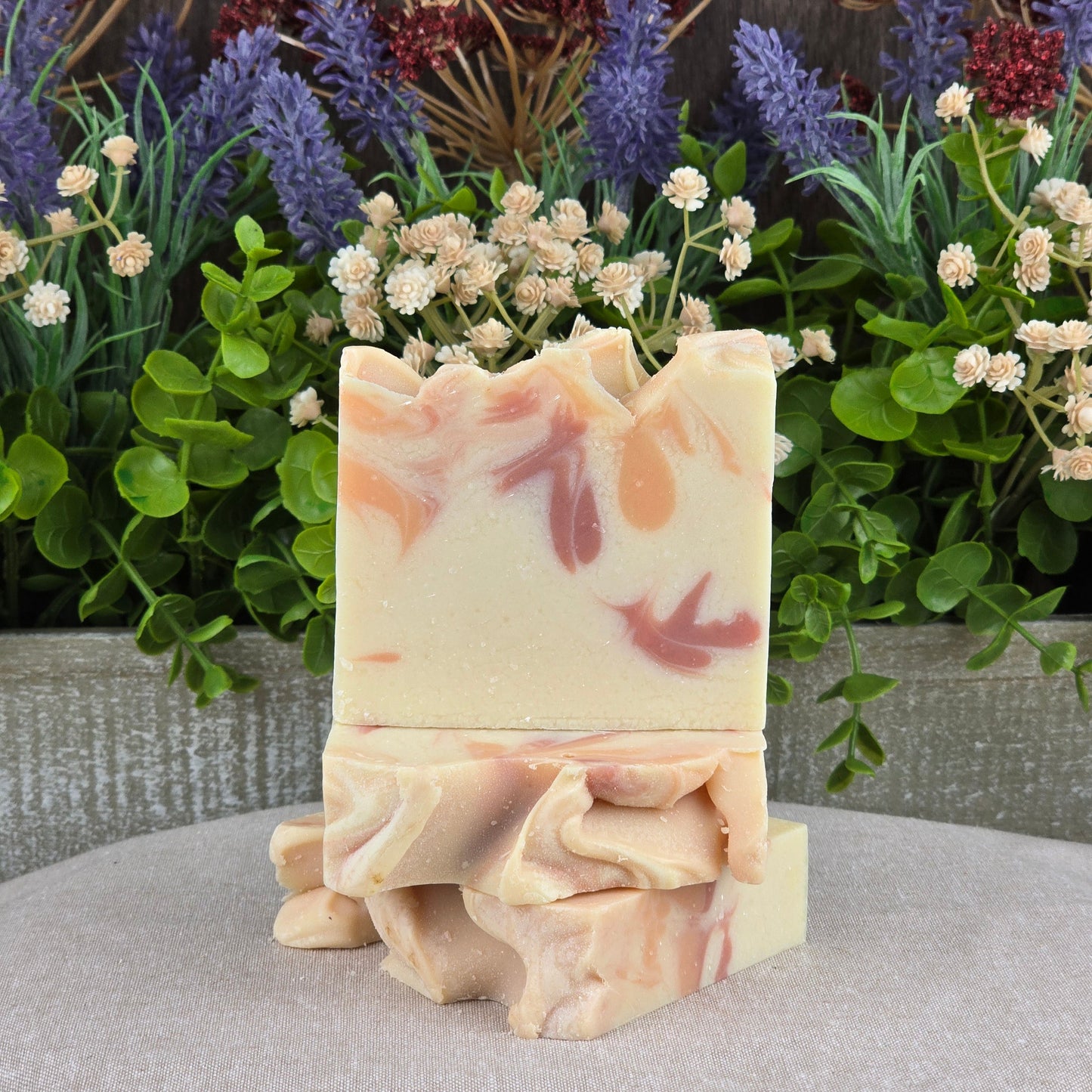 Two Apple + Spice Handmade Coconut Milk Artisan Soap bars by Jarhead Aromatics, featuring swirled pink and orange patterns, are stacked on a beige surface with green leaves and purple and white flowers in the background.