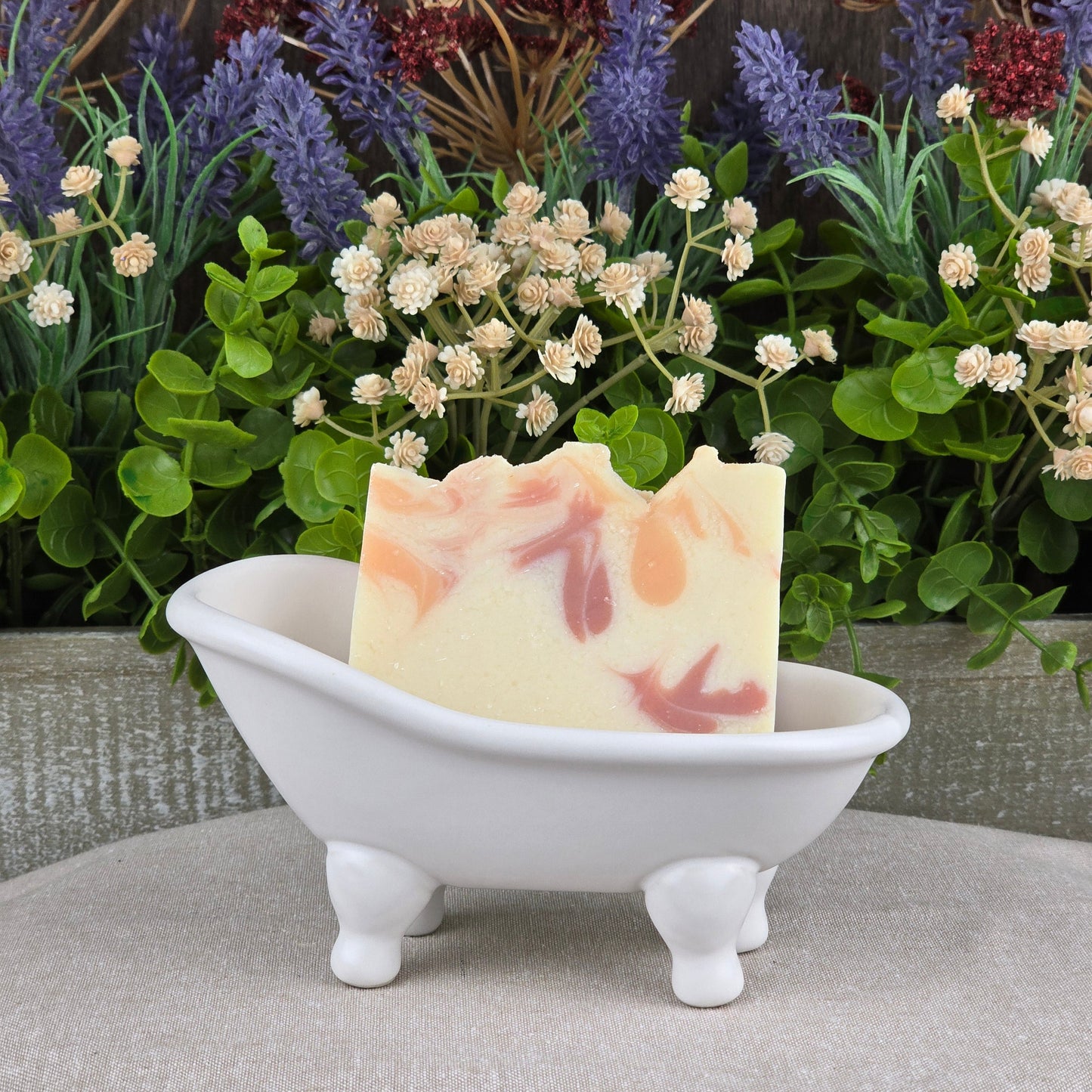 A decorative bar of Jarhead Aromaticsโ Apple + Spice Handmade Coconut Milk Artisan Soap with a marbled pattern sits in a white clawfoot bathtub dish, accented by green leaves and purple and white artificial flowers.
