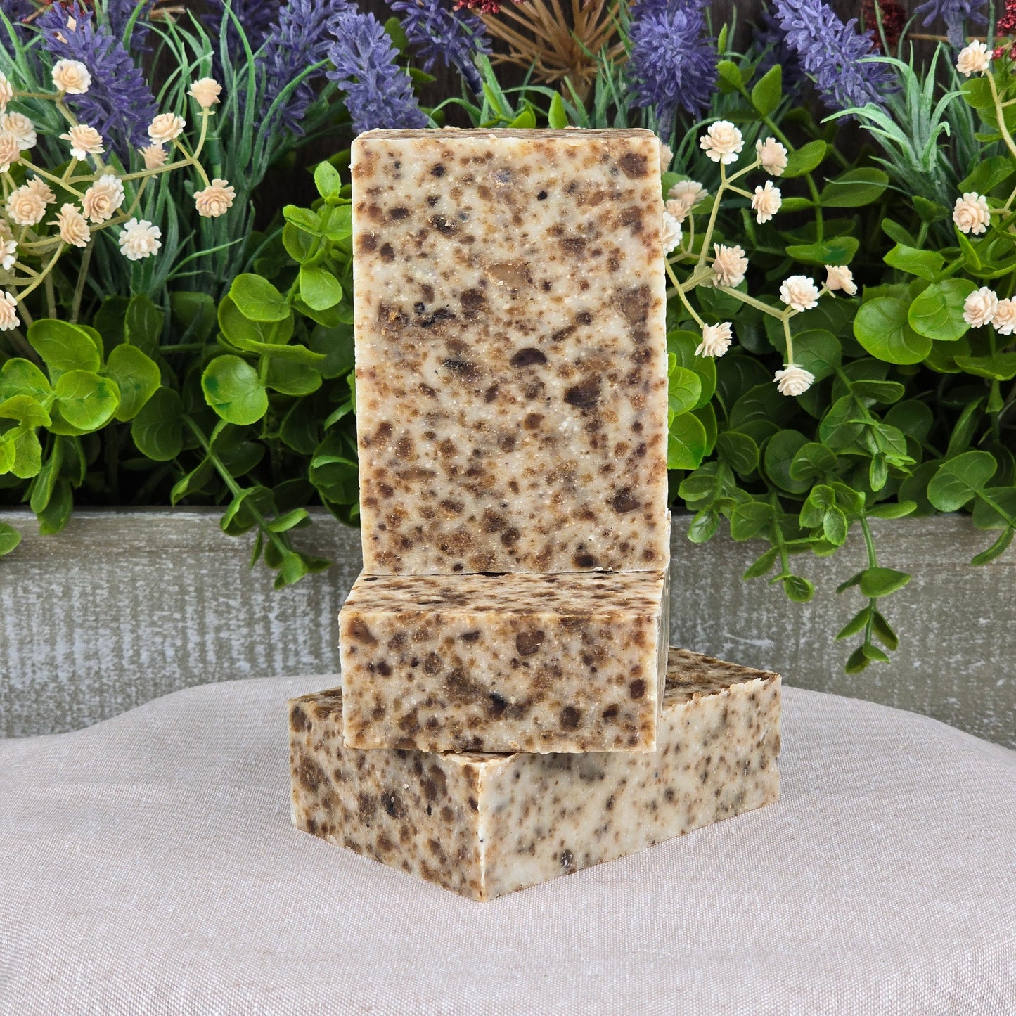 Three rectangular bars of Jarhead Aromatics Black Soap + Lavender Handmade Artisan Soap, all-natural with a speckled appearance, are stacked in front of green foliage and small flowers on a light-colored fabric surface.