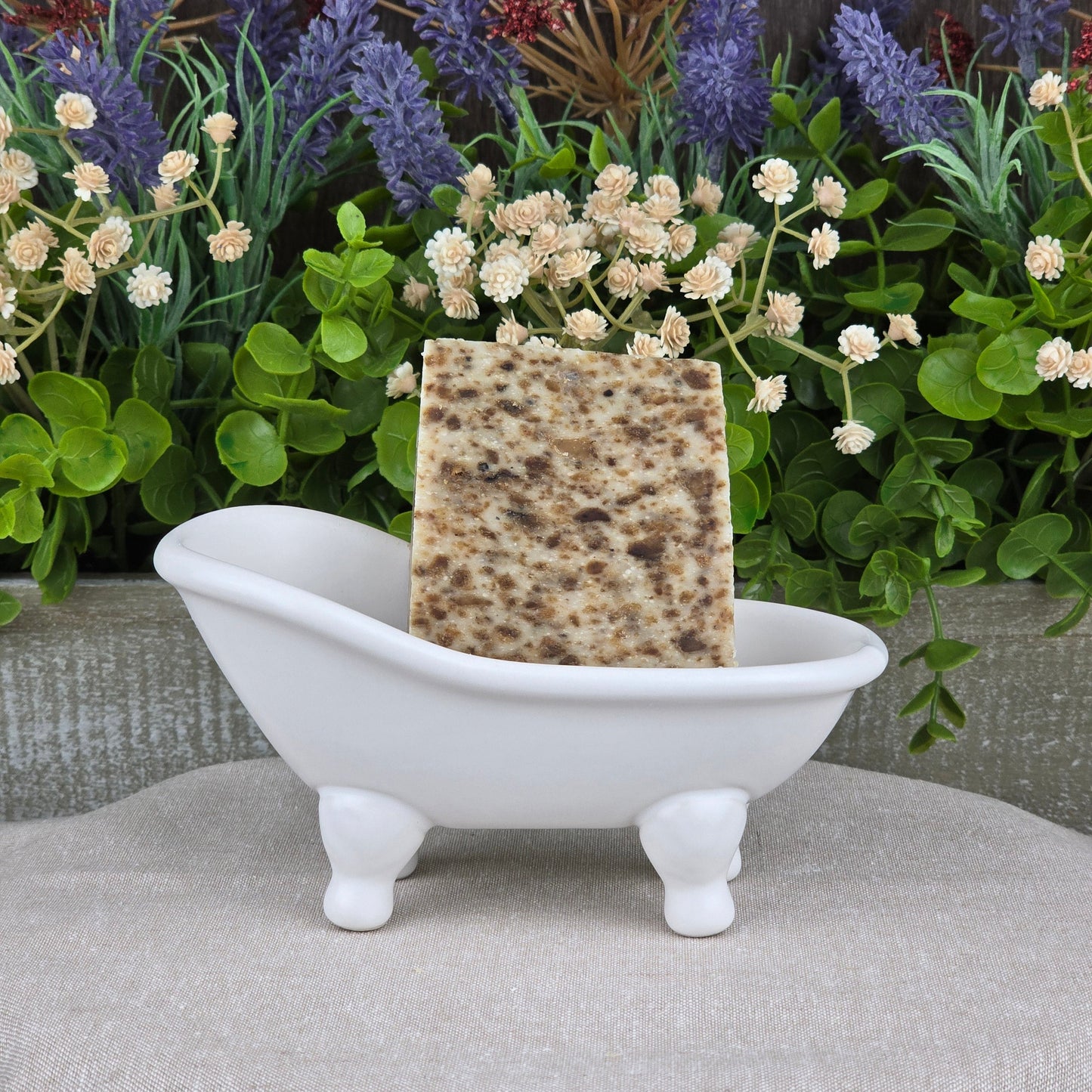 A rectangular, speckled bar of Black Soap + Lavender Handmade Artisan Soap by Jarhead Aromatics rests in a white clawfoot bathtub soap dish, with green leaves and colorful flowers in the background.