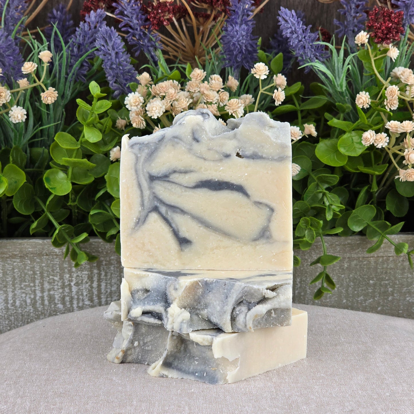 Cashmere + Santal Handmade Coconut Milk Artisan Soap by Jarhead Aromatics features a cream and gray marbled design, stacked among green foliage and purple and cream flowers on neutral fabricโbringing natural charm to every bar.